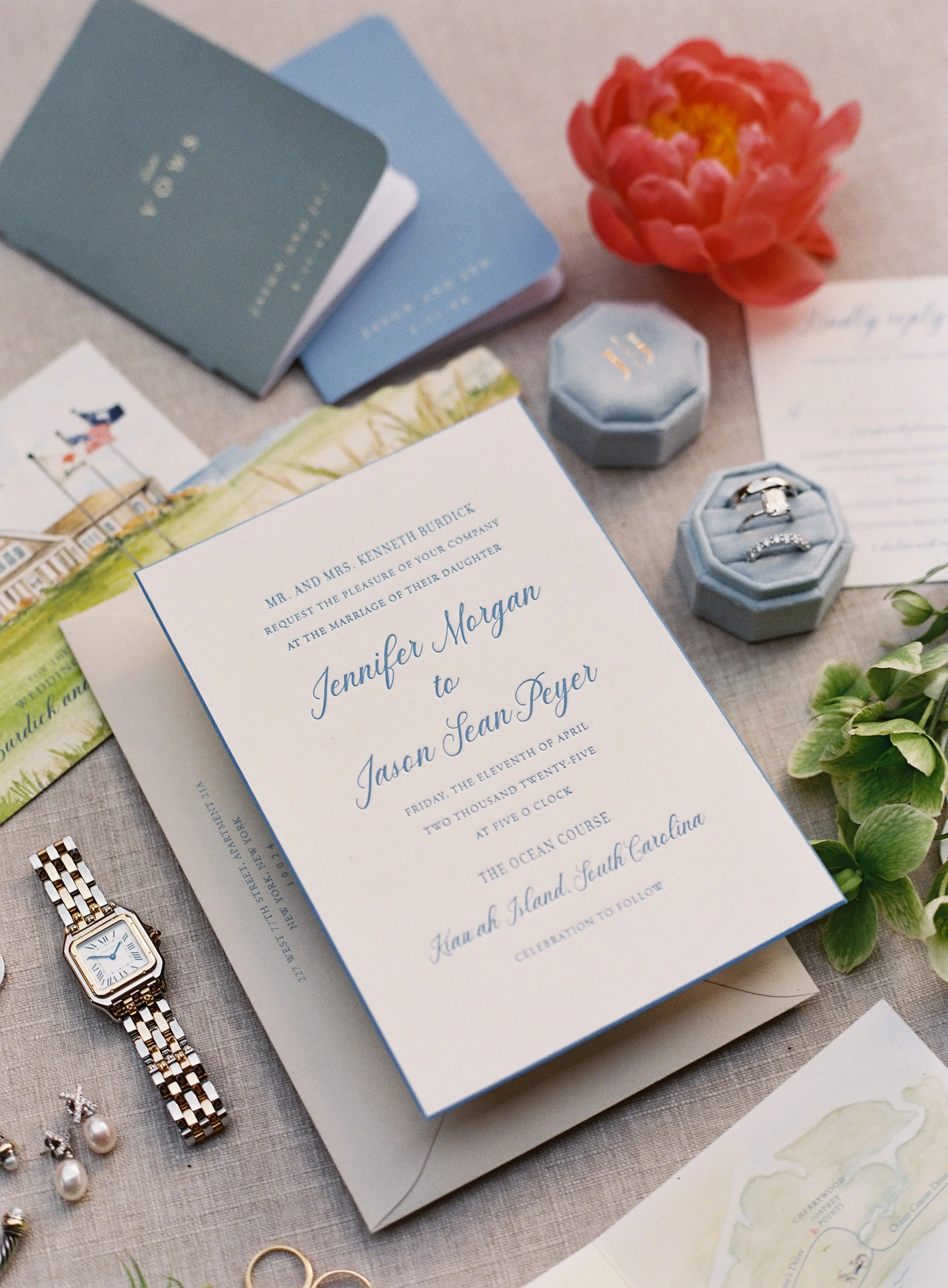 Wedding invitation surrounded by jewelry, floral arrangements, and rings on a table.