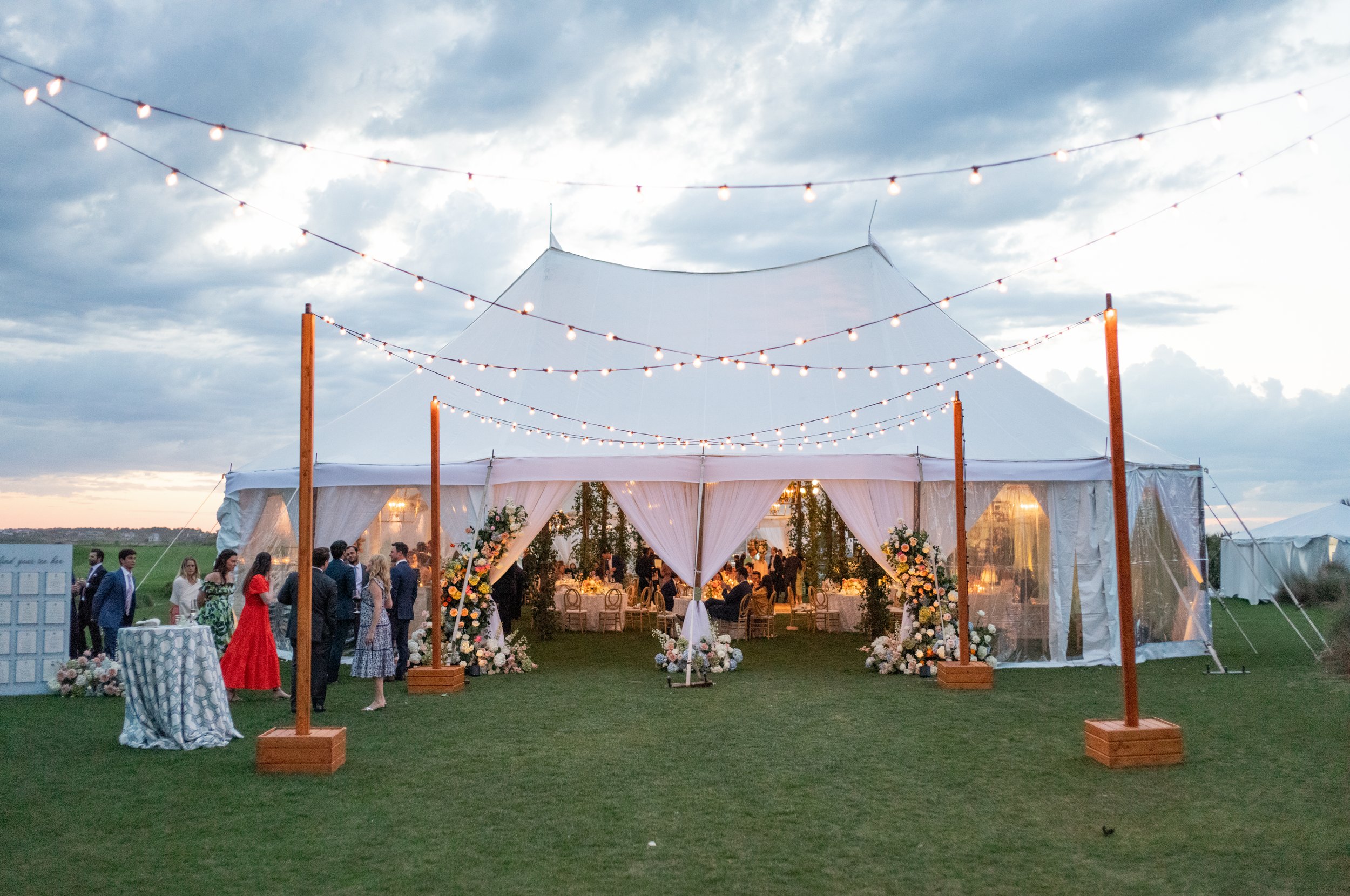 Outdoor wedding reception under a large white tent with string lights, floral decorations, and guests mingling on the grass at sunset.