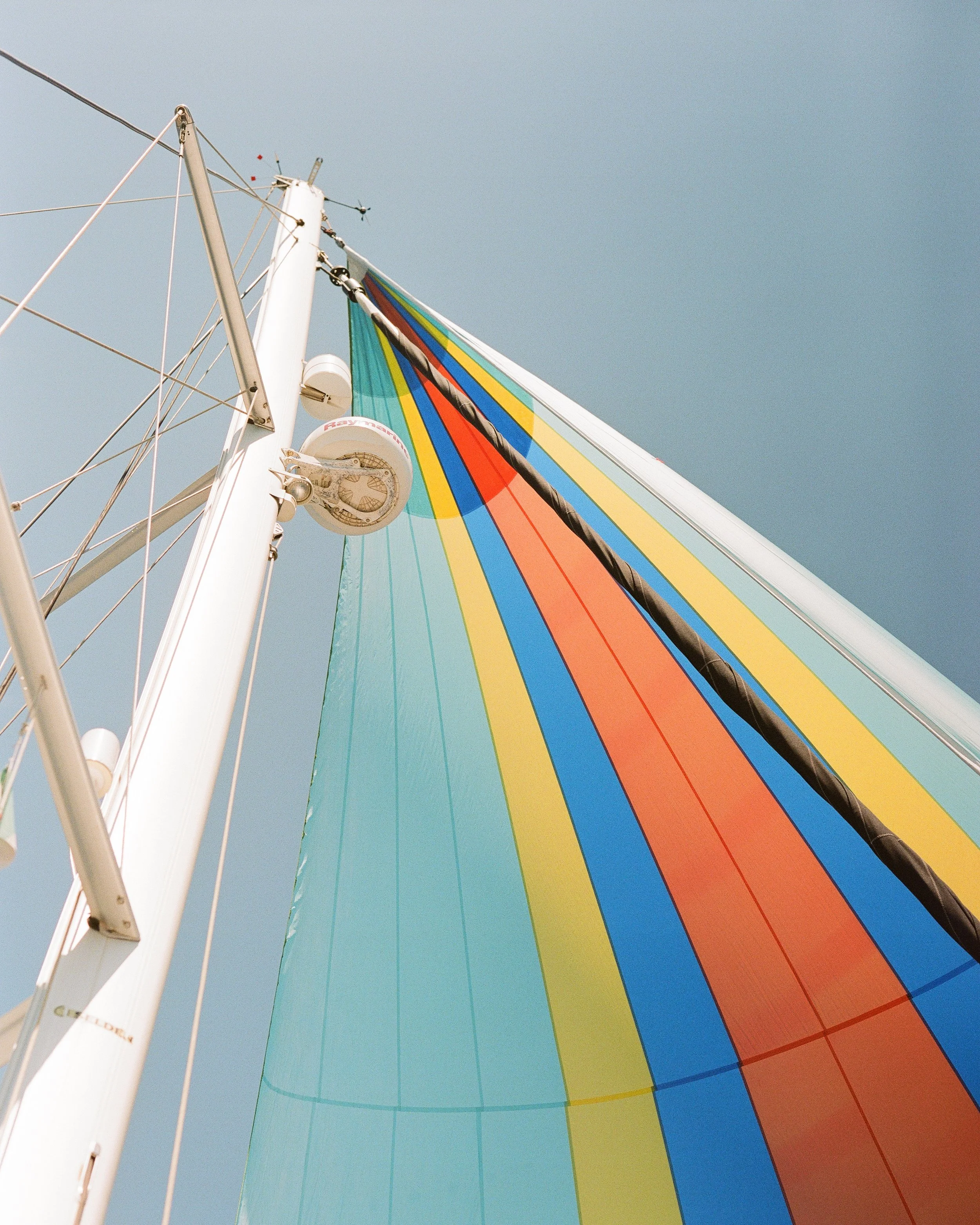 Looking up at a sailboat's mast in Sorrento, Italy with a vibrant, multi-colored sail, against a clear blue sky.