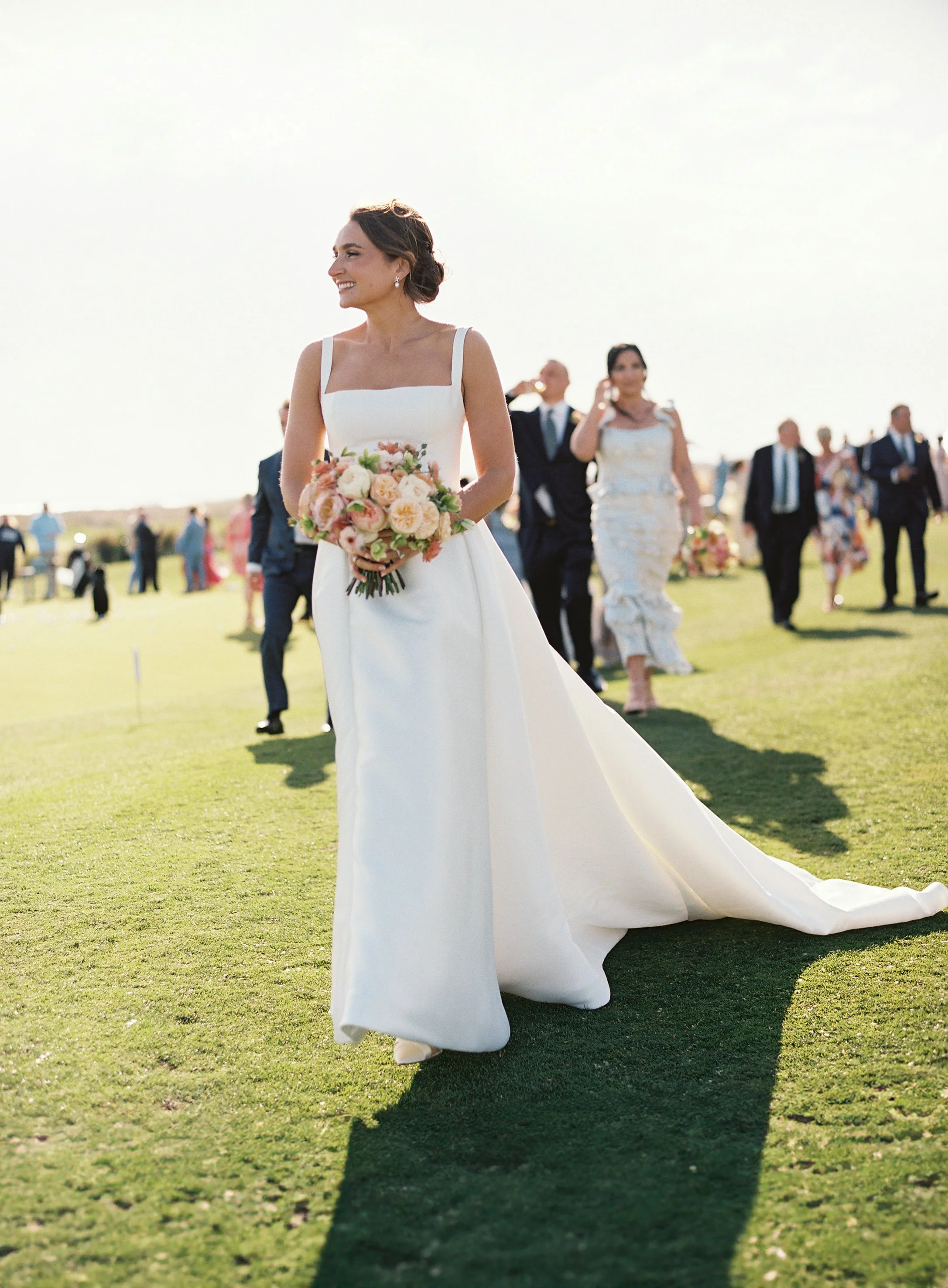 Bride in a white wedding dress holding a bouquet of flowers walking on a grassy field during a wedding celebration.