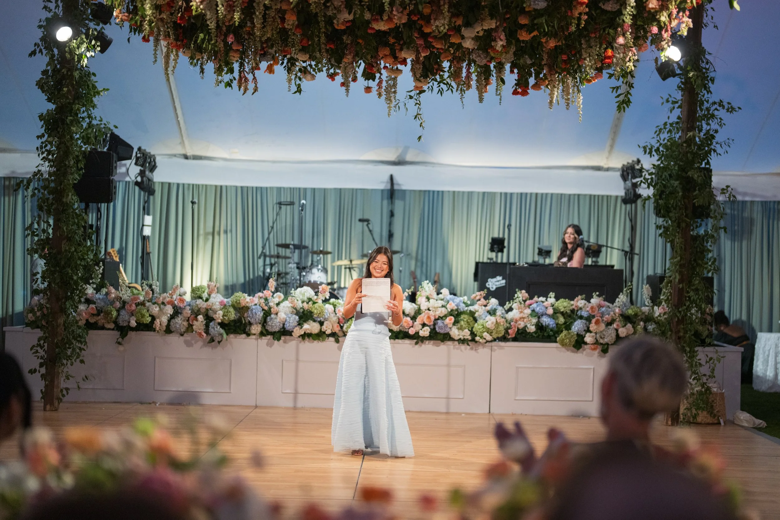 A woman in a light blue dress holding a piece of paper, smiling on a wooden dance floor at a wedding reception. Behind her is a stage decorated with pink, white, and purple flowers, with a drummer and a DJ or musician in the background. The ceiling i