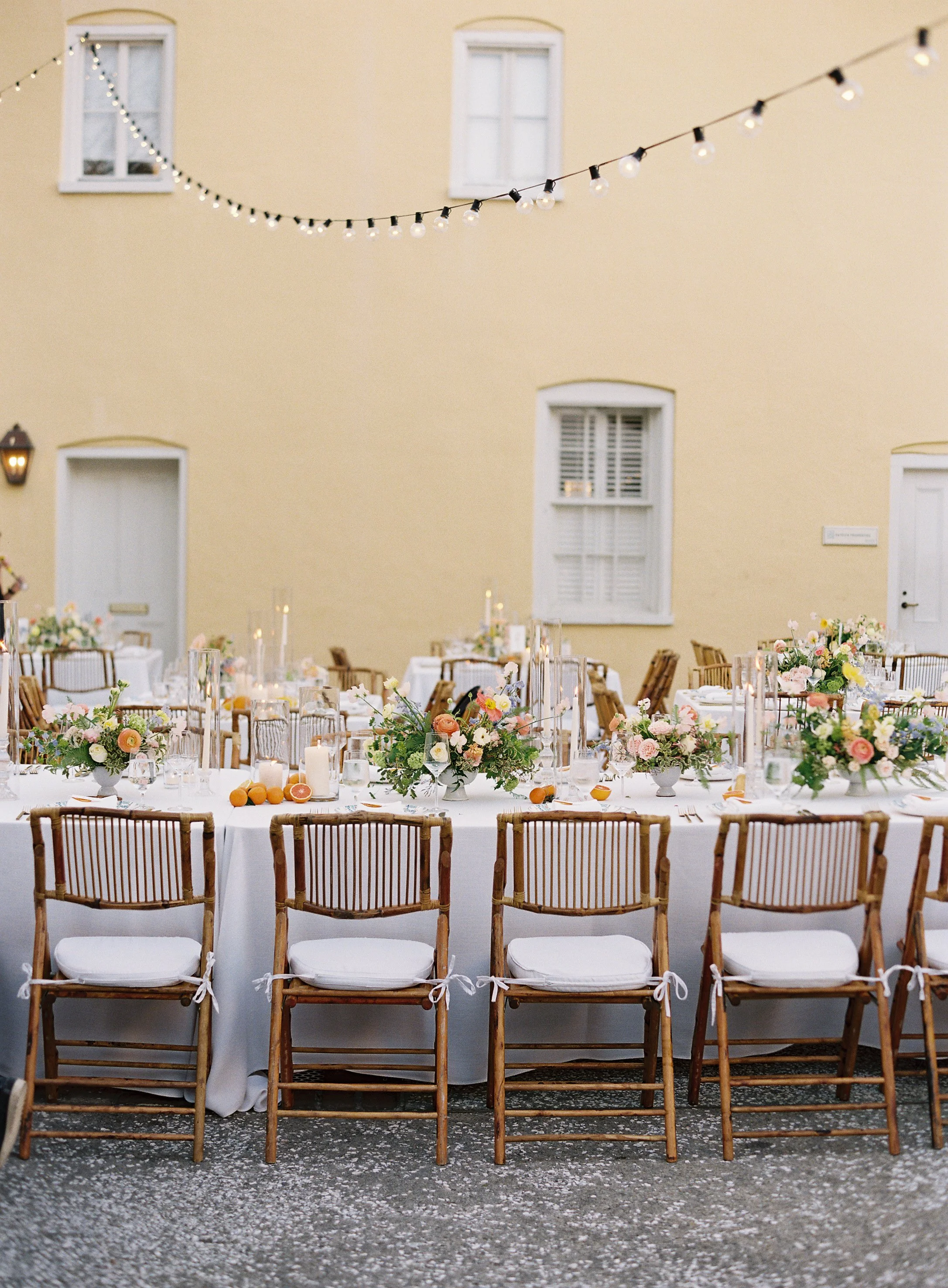 Elegant outdoor wedding reception table setting with floral centerpieces, candles, and string lights against a yellow wall.