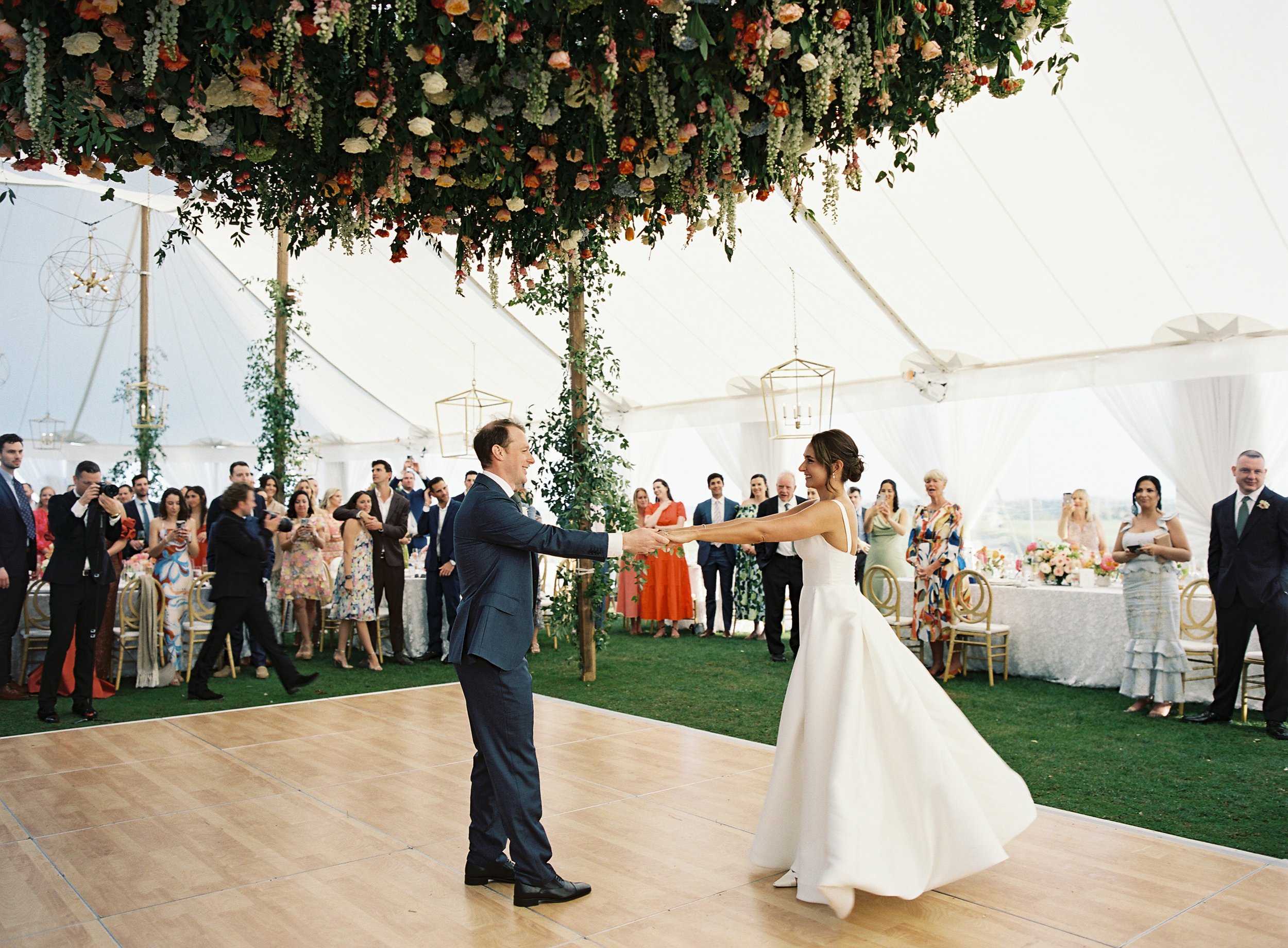A bride and groom are dancing at their wedding reception at the Ocean Course on Kiawah Island, inside a white sailcloth tent. The tent has hanging lanterns and a large floral arrangement above the dance floor.