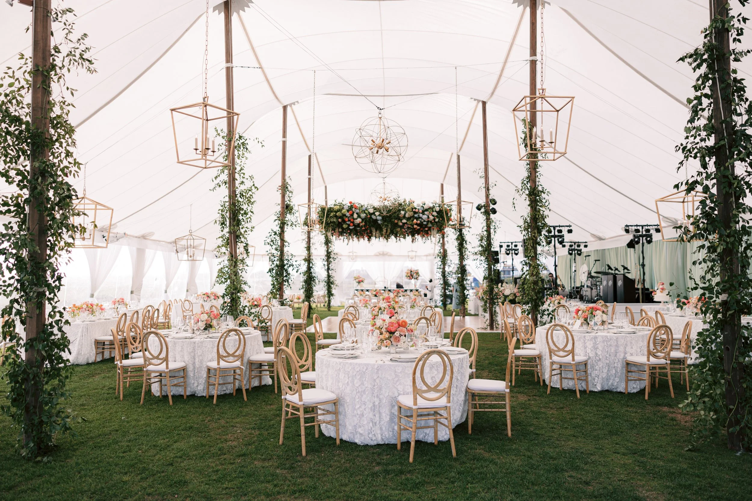A decorated outdoor wedding or event tent with round tables covered in white lace tablecloths, surrounded by wooden chairs with white cushions. The tables have floral centerpieces with pink, peach, and white flowers. Hanging from the ceiling are geom