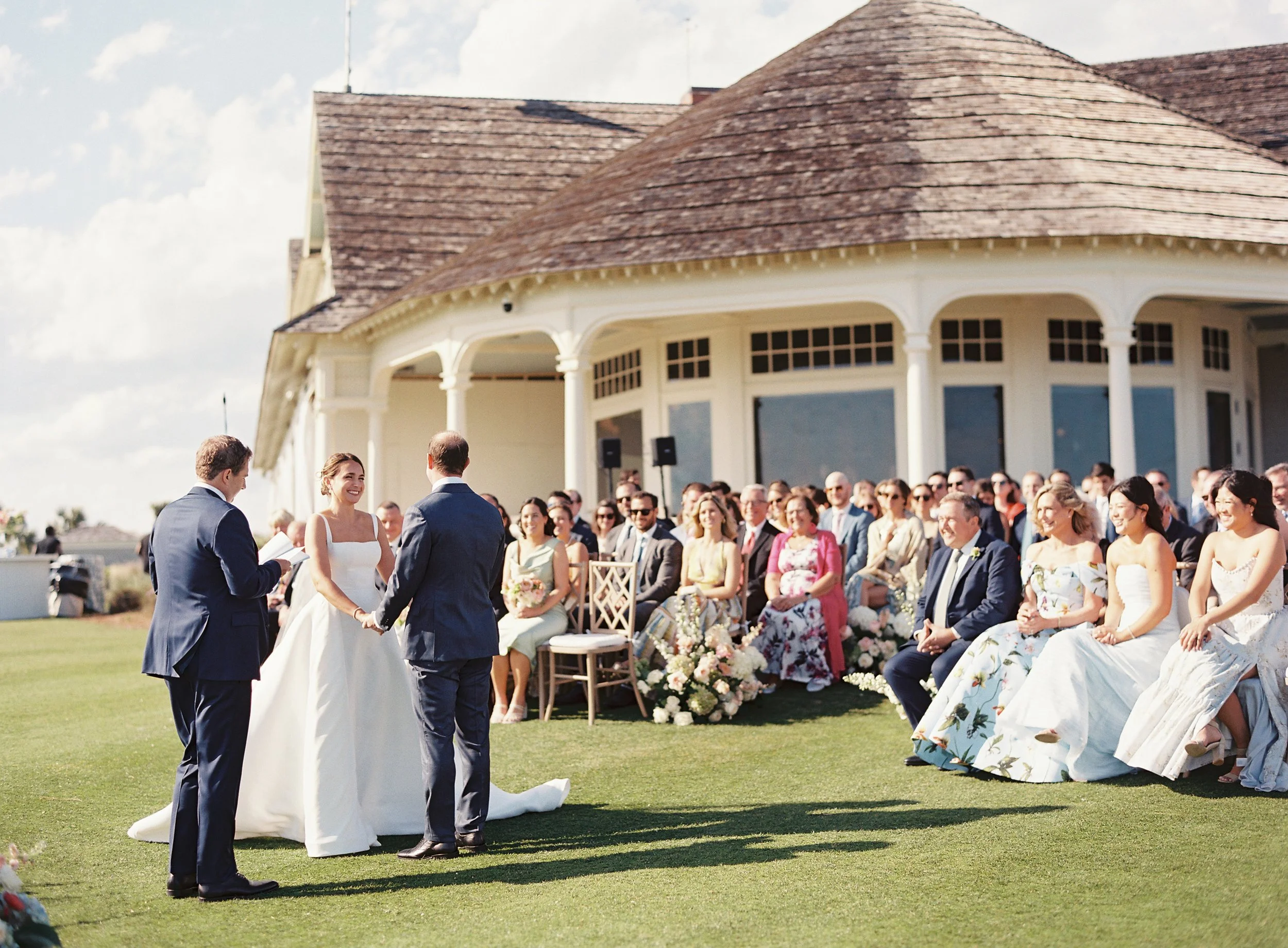 Black-haired woman in a strapless white wedding dress holding hands with a man in a navy suit during an outdoor wedding ceremony. An officiant holds a book, officiating the ceremony. Guests sit on chairs and in the front row, smiling. The background 