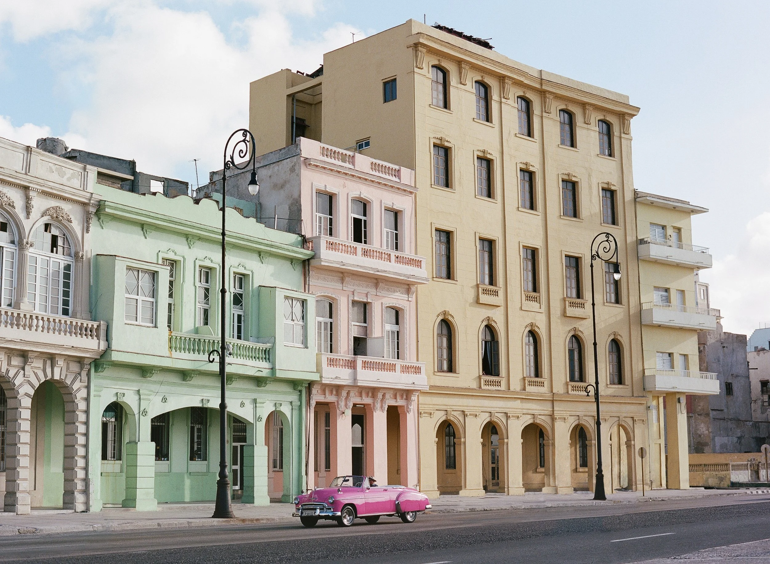 Colorful vintage buildings in Cuba, lining a city street with a pink classic car parked in front and two black street lamps, under a partly cloudy sky.