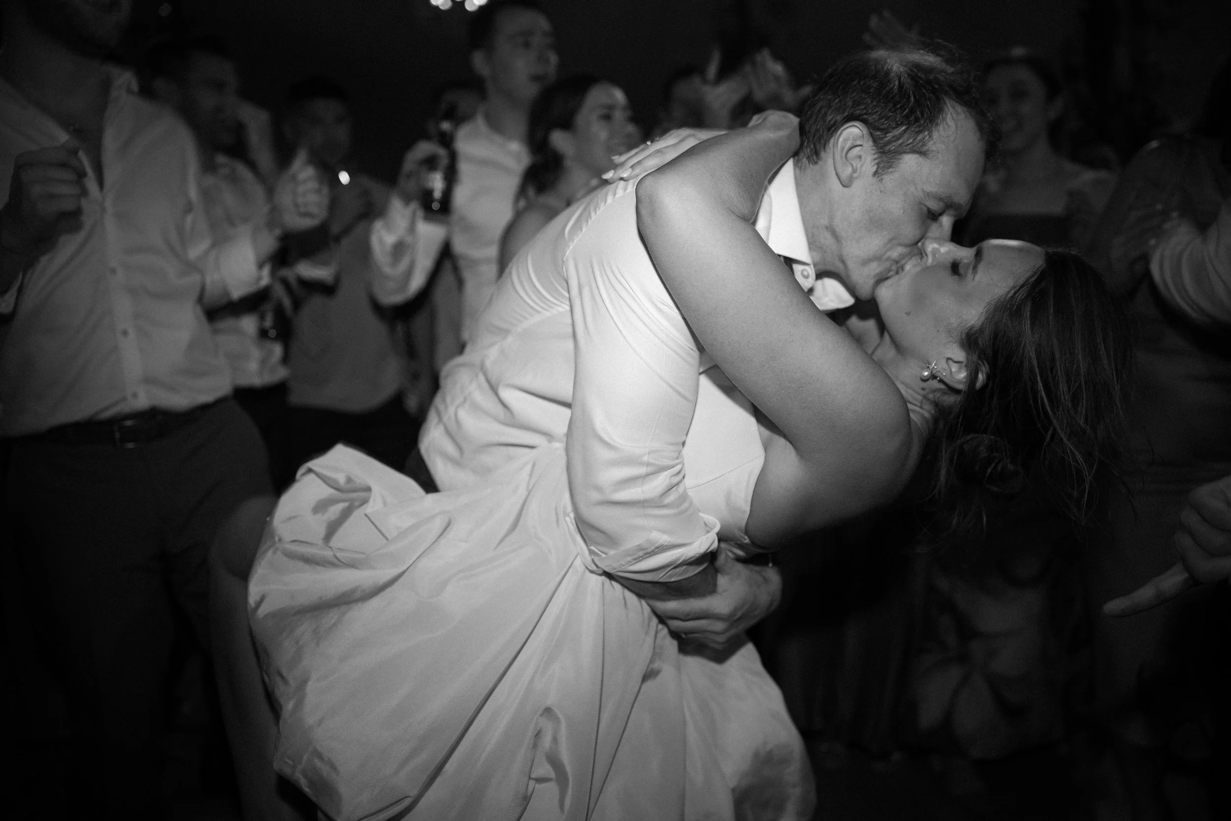 A man and woman sharing a kiss, with the man dipping the woman, at a crowded party or celebration.