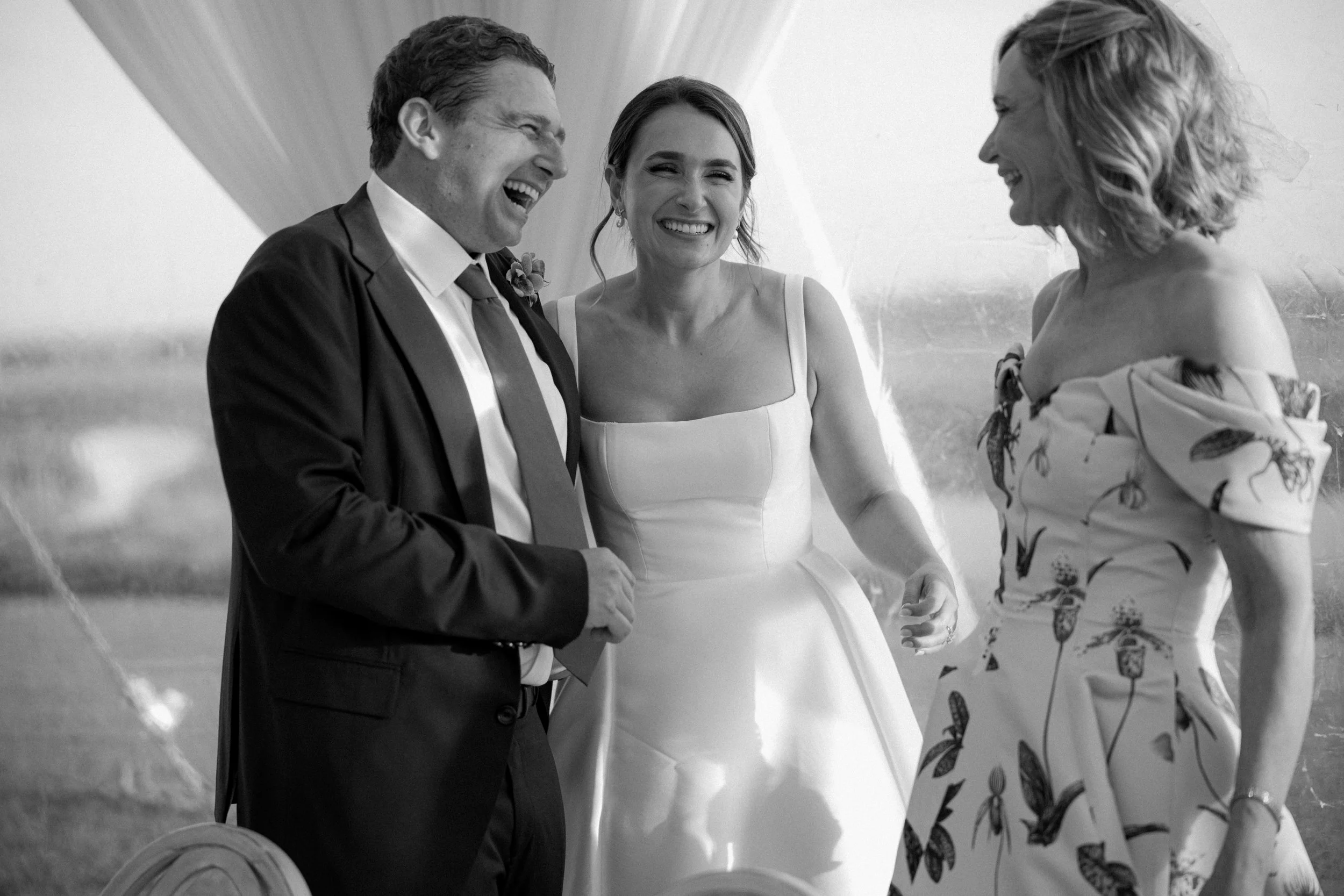Black and white photo of a wedding scene with three people smiling and laughing, the bride in a wedding dress, the groom in a suit, and a woman in a floral dress.
