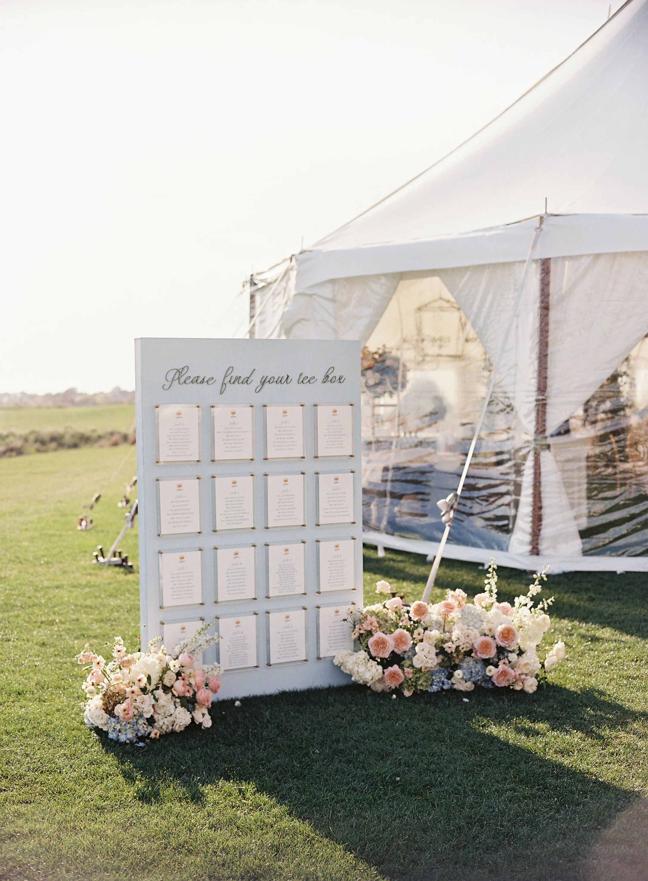 A white seating chart display with multiple paper cards, decorated with pink and white flowers, set outside near a white tent.