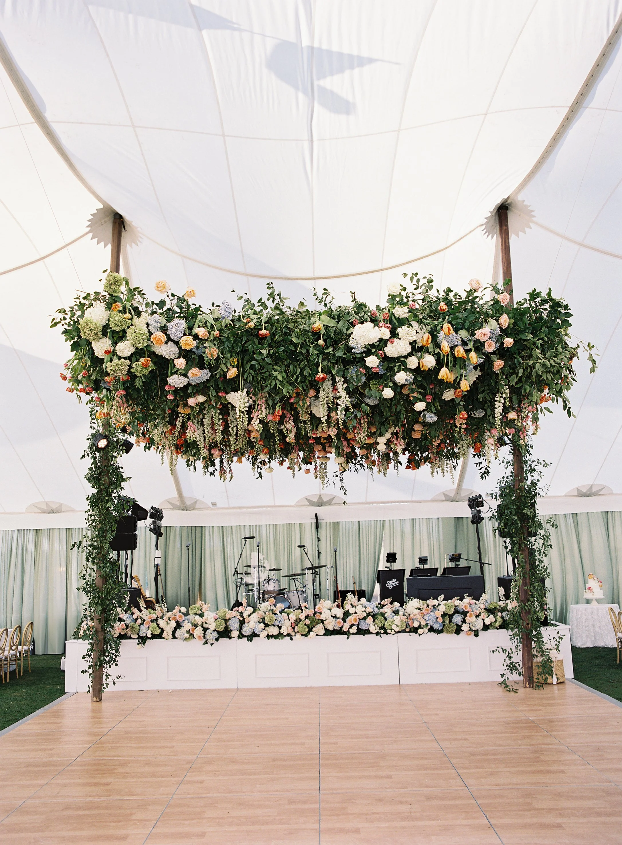 Decorative wedding stage with a hanging floral arrangement and musical instruments underneath, inside a large white tent.