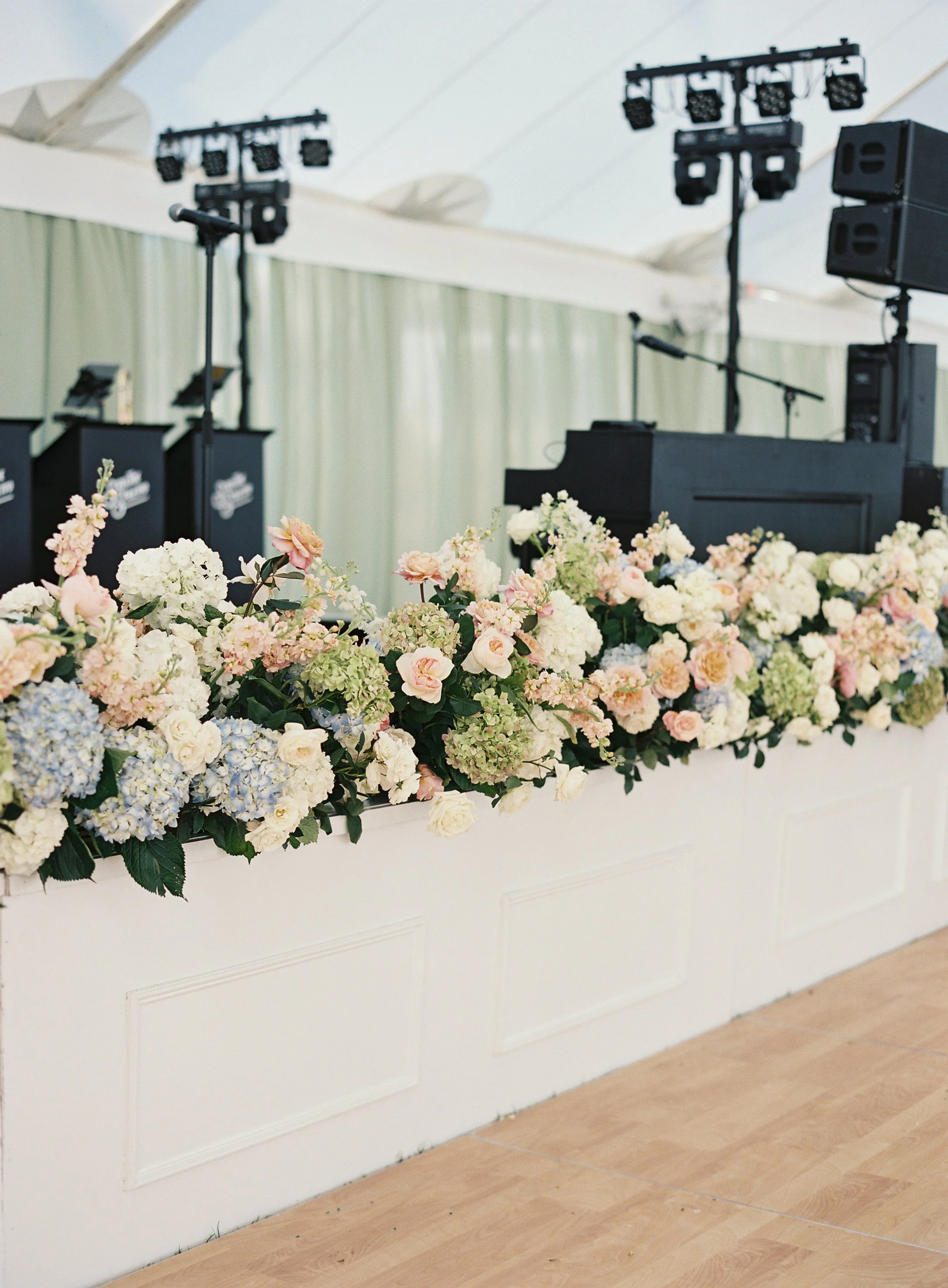 A stage decorated with a large floral arrangement of white, pink, and green flowers, with musical equipment and lights in the background, set up in a white tent for an event.