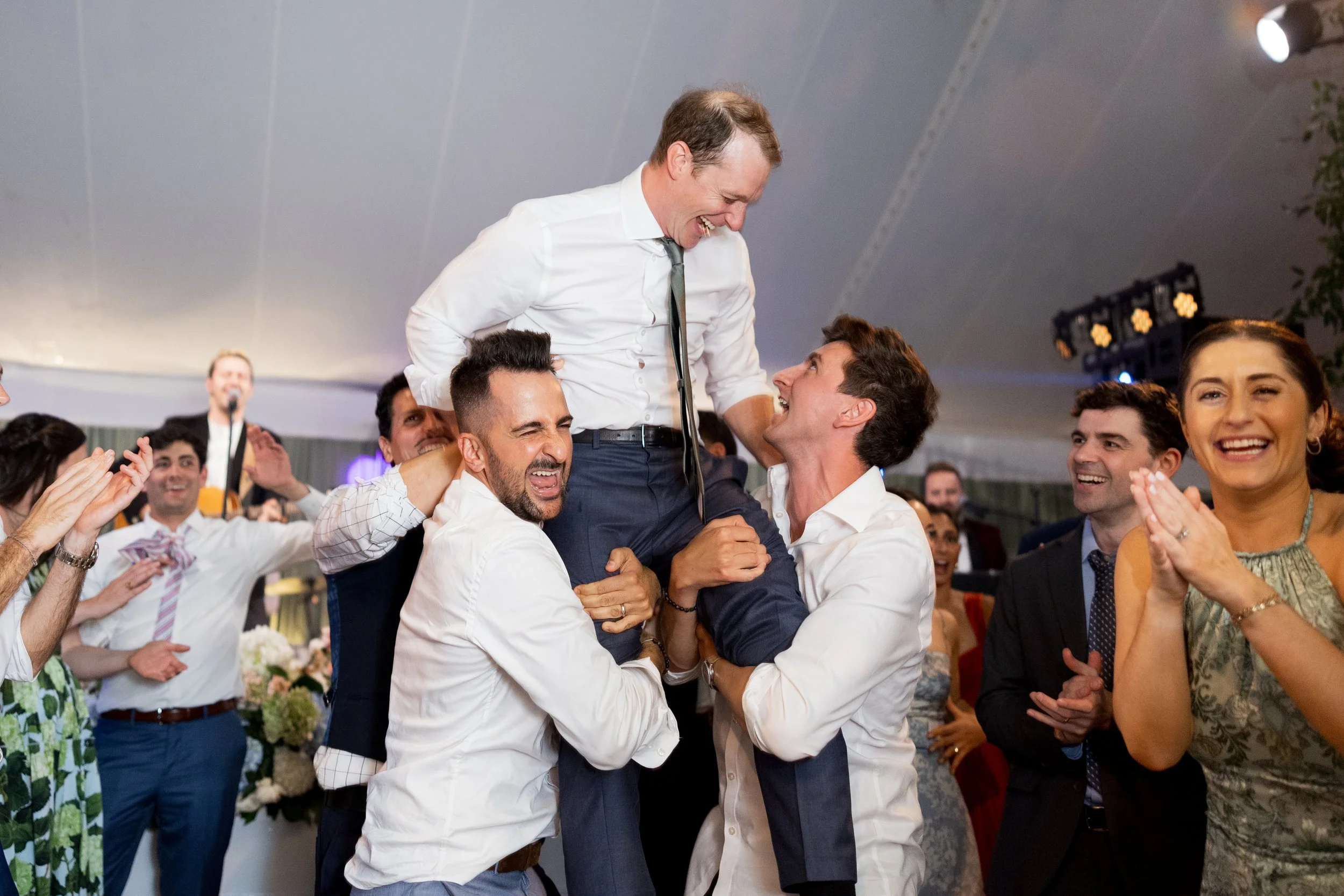 Group of people celebrating at a wedding, lifting the groom in the air, with smiles and applause.
