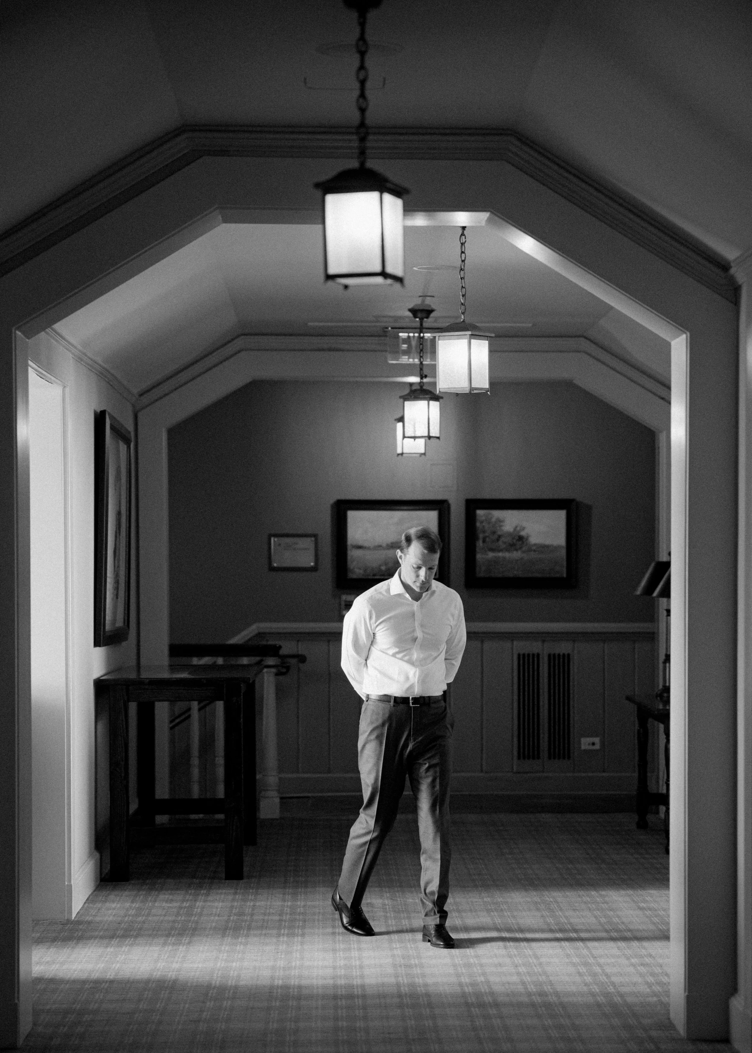 A man in a white shirt and dark pants, walking with his head bowed in a hallway with hanging ceiling lights and framed pictures on the wall.