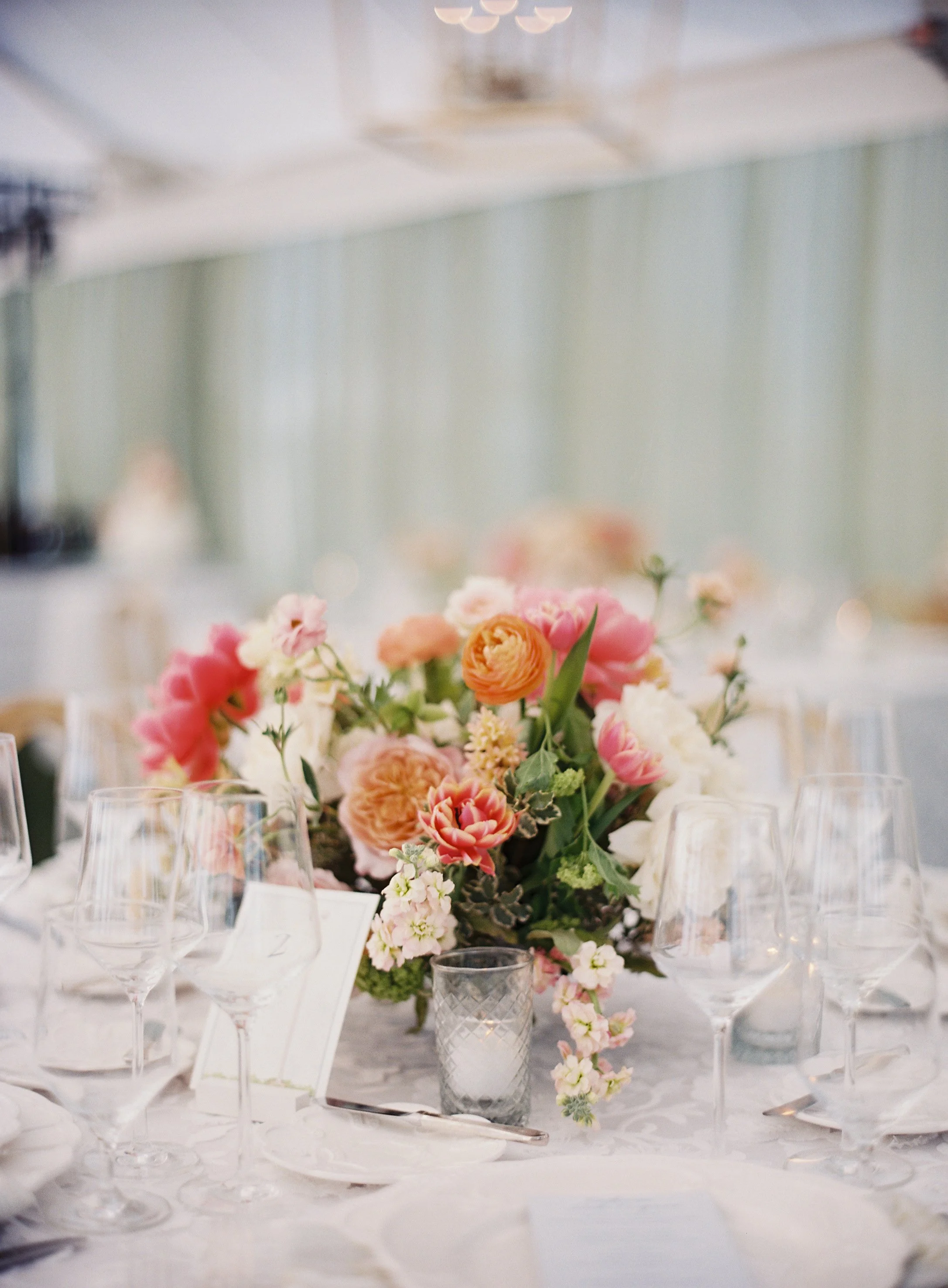 Elegant table centerpiece with pink, peach, and white flowers surrounded by wine glasses and a small candle holder, set in a well-lit event hall.