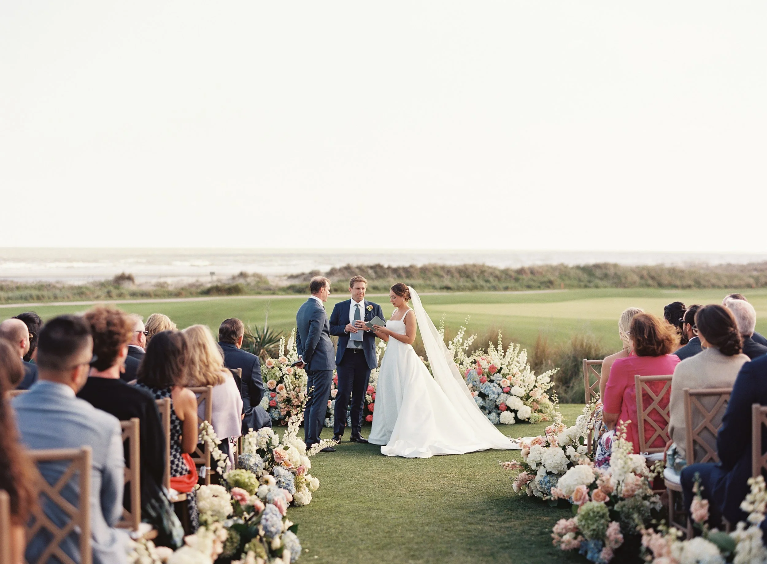 A wedding ceremony taking place outdoors on a grassy area, with a view of the ocean in the distance. The bride and groom stand in front of an officiant, surrounded by floral arrangements and seated guests.