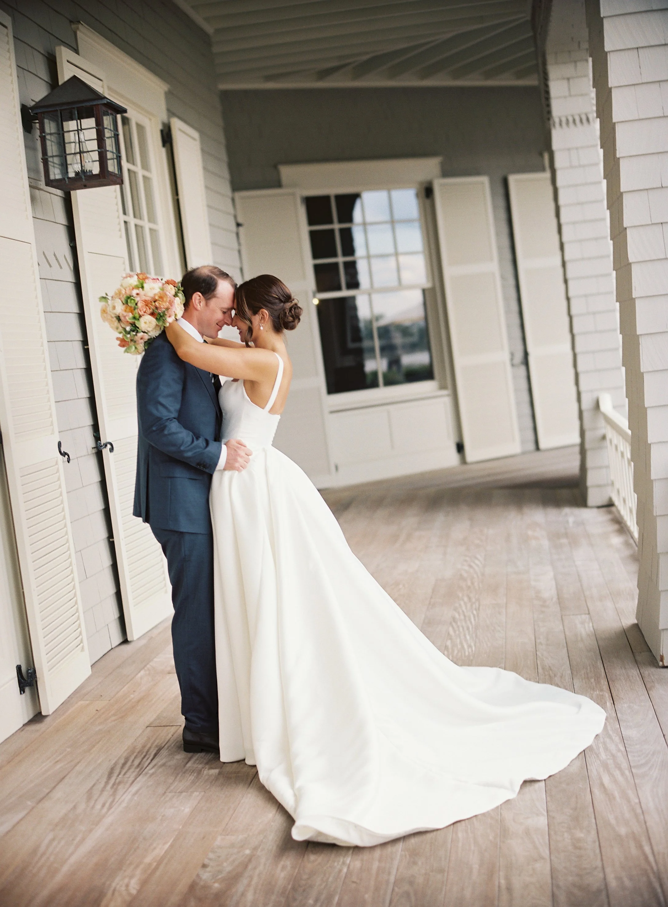 A bride and groom embrace on a porch, with the bride holding a bouquet of flowers, both smiling with noses touching, in a wedding setting.