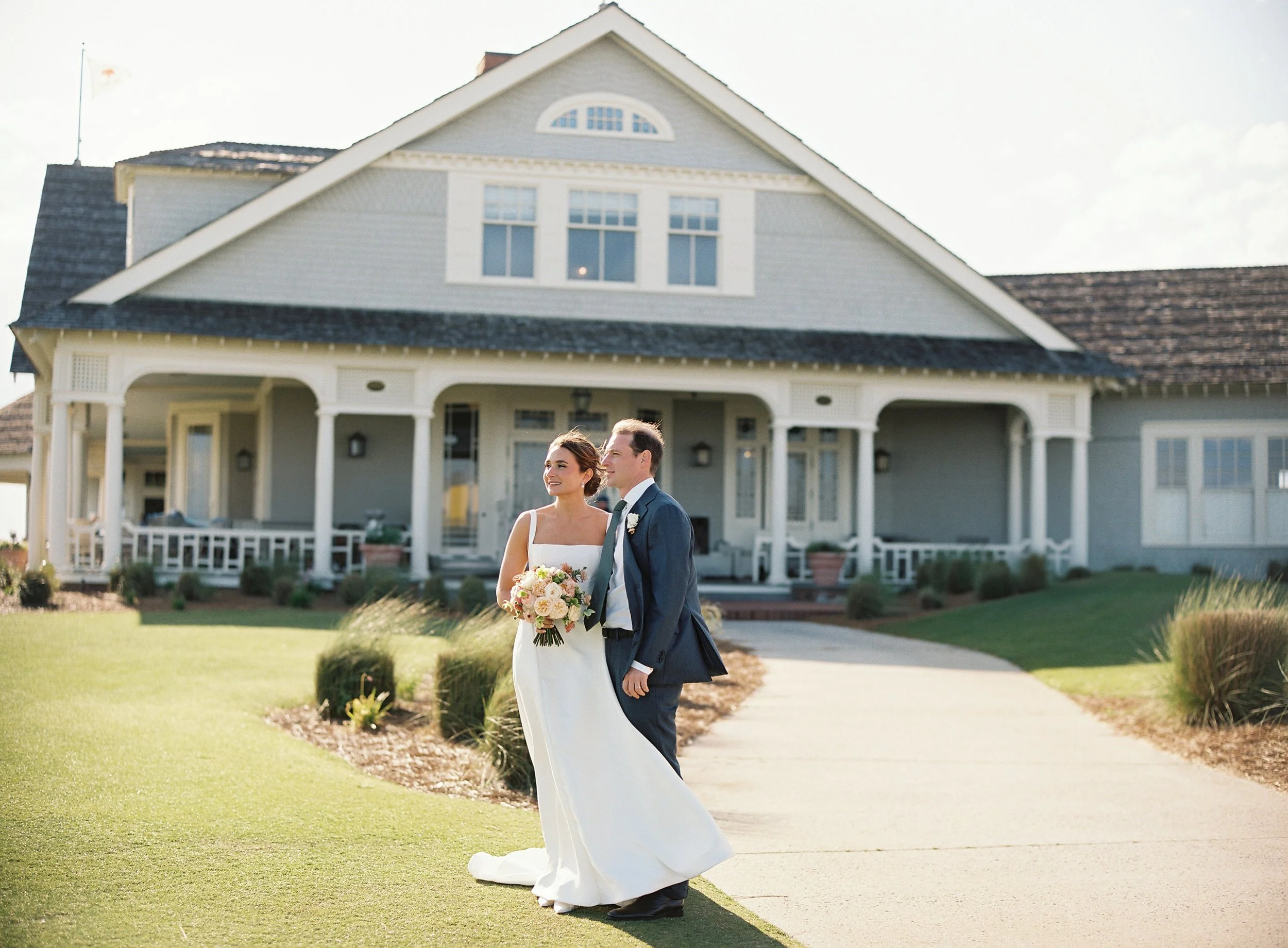 A bride and groom standing outside a large house, smiling and holding a bouquet of flowers, during a wedding photoshoot.
