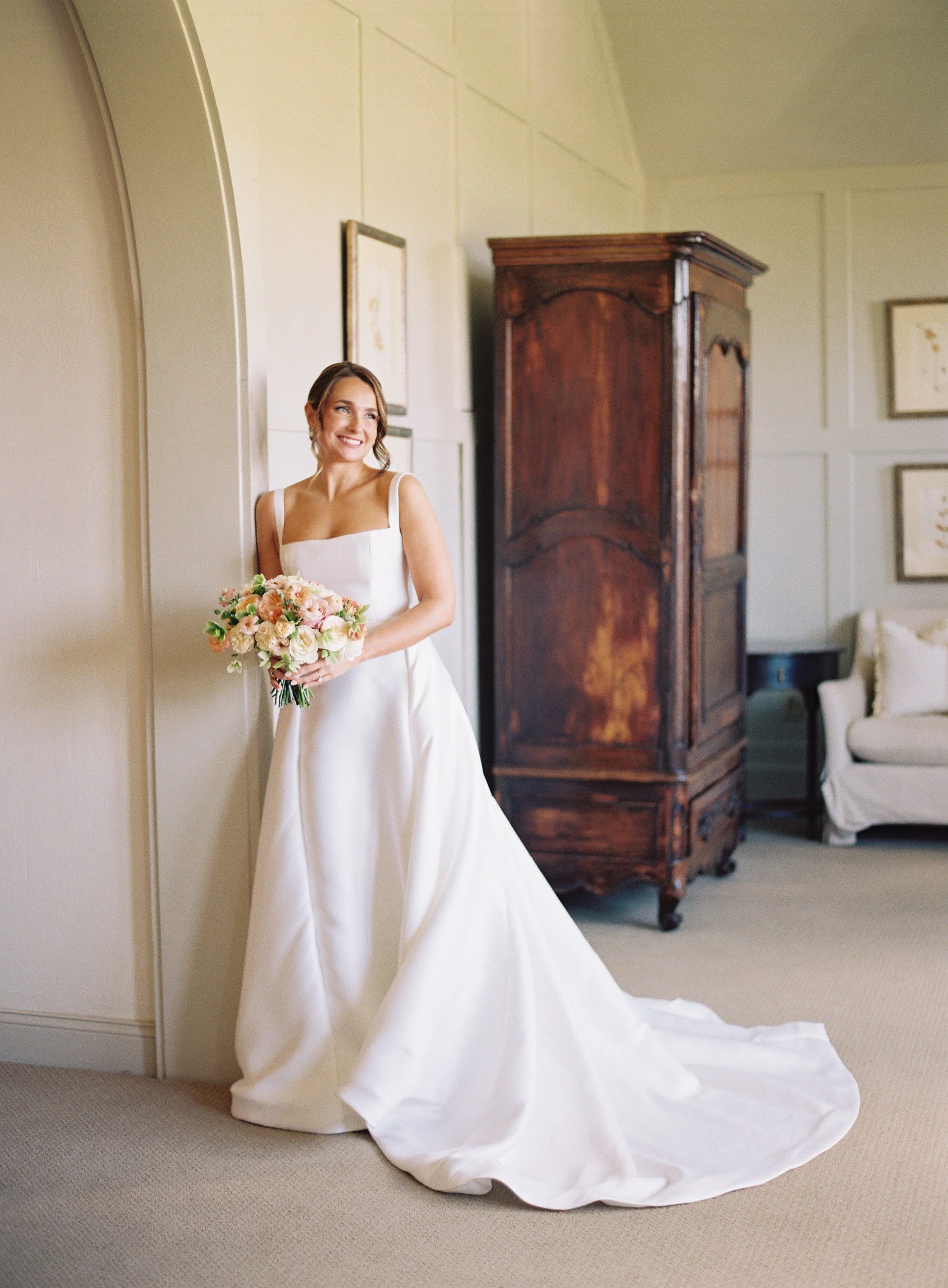 Bride in white wedding dress holding a bouquet, standing in a room with a vintage wooden armoire and a beige sofa.