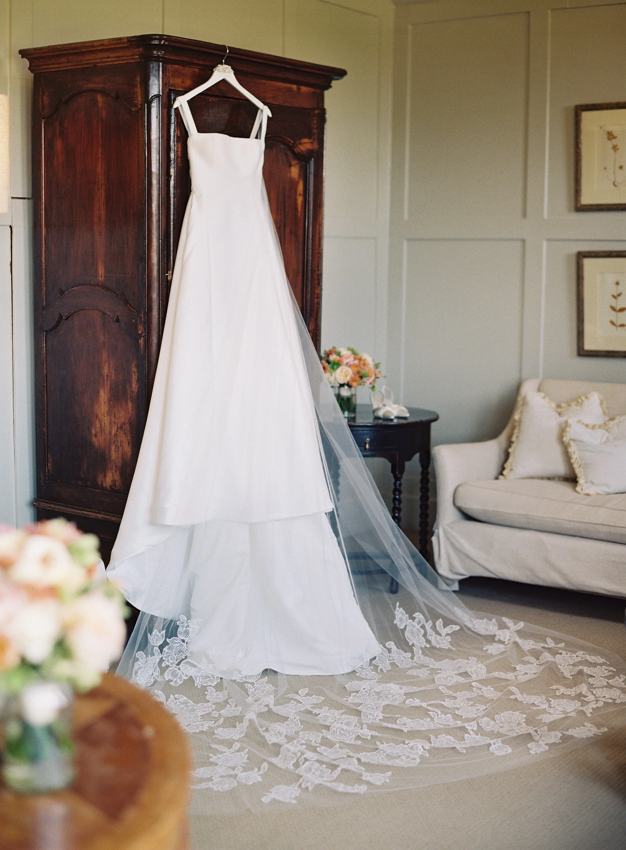 A white wedding dress hanging on a hanger on a wooden armoire in a cozy room with light-colored paneled walls, a beige carpet, a small dark side table with a bouquet of pink and peach flowers, and a cream-colored sofa with plush pillows.