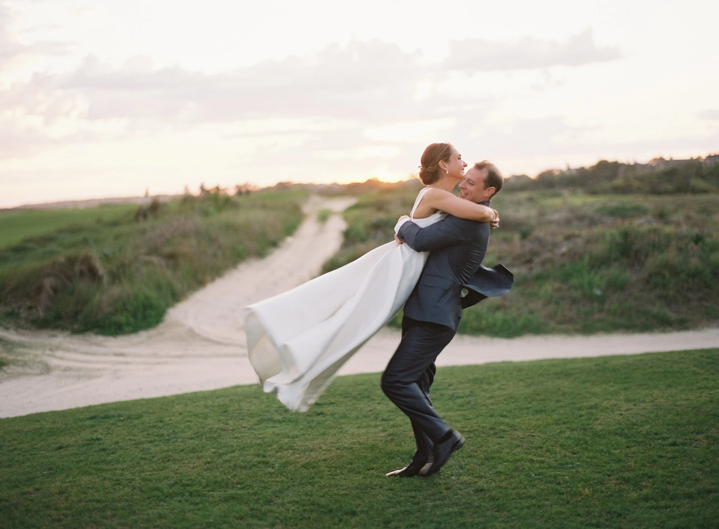 A groom in a dark suit is lifting a bride in a white wedding dress in a grassy outdoor area at sunset. The couple is smiling and sharing a joyful moment on a path with green hills in the background.