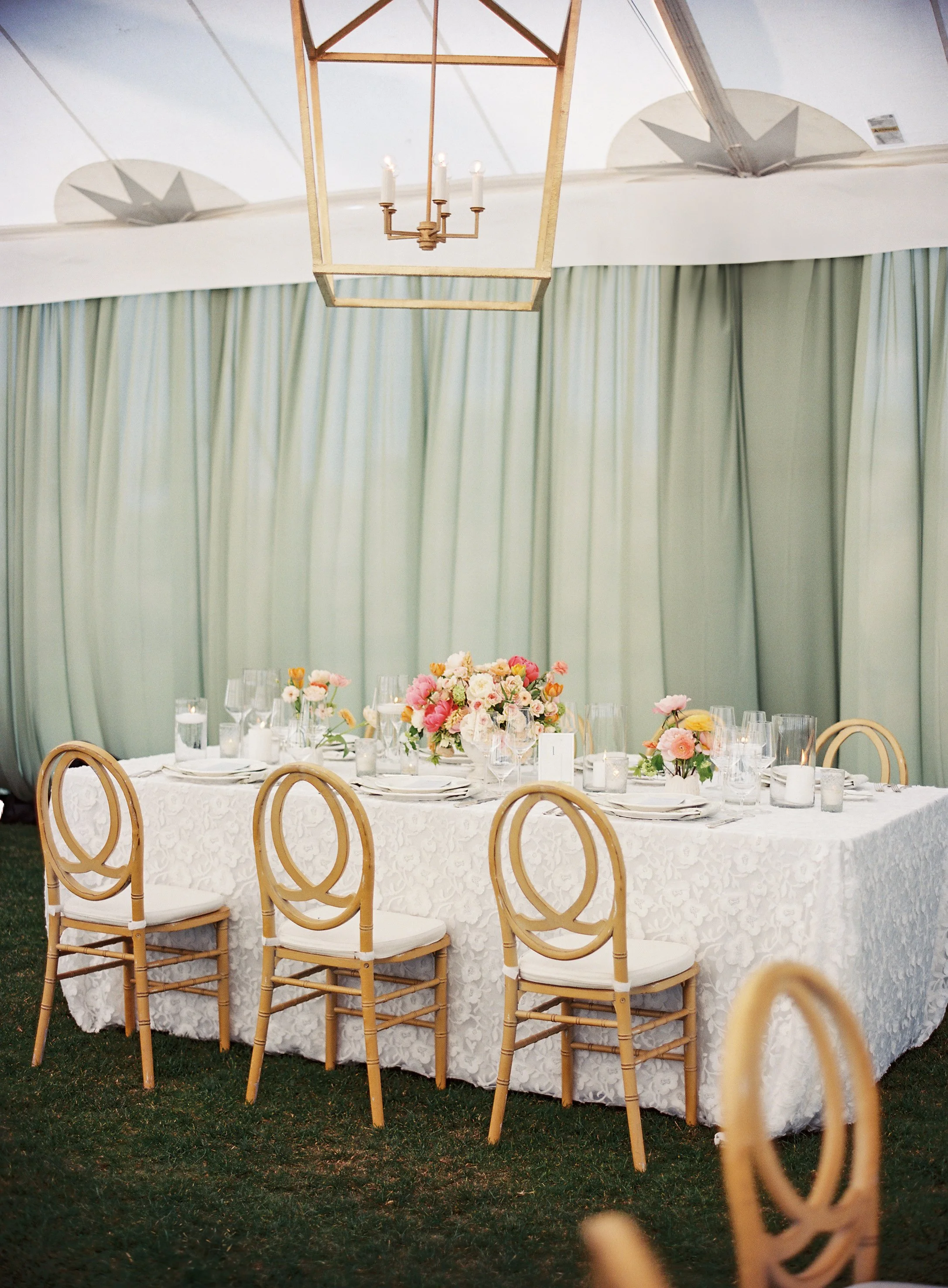 Elegant outdoor dining table setup with a white lace tablecloth, centerpieces of pink and white flowers, glassware, and candles, under a tent with green curtains and a wooden chandelier.