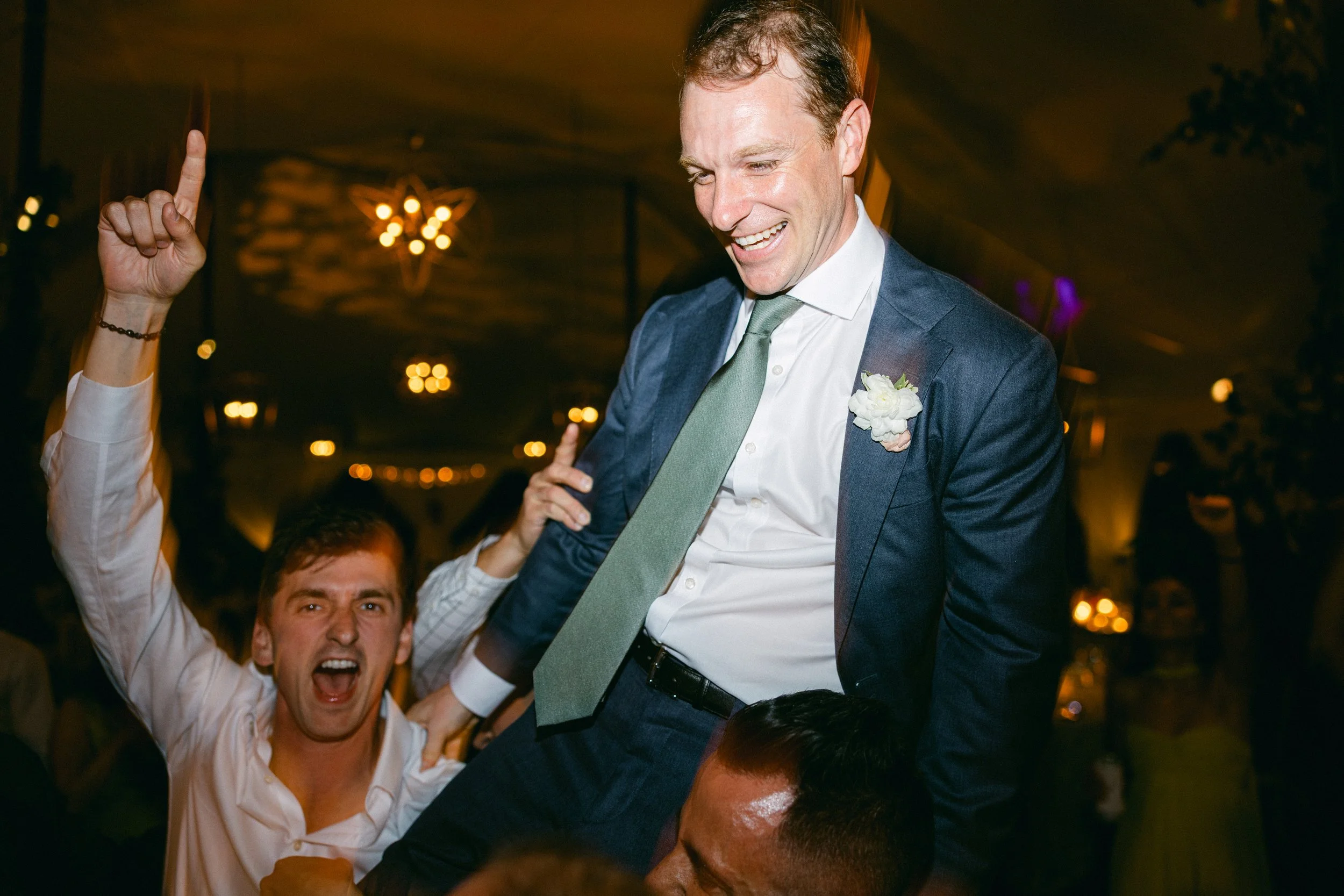A man in a blue suit and light green tie sitting on someone's shoulders at a celebration, smiling and pointing upward. A man in a white shirt behind him is cheering with his mouth open, and there are warm lights and a decorated indoor venue in the ba