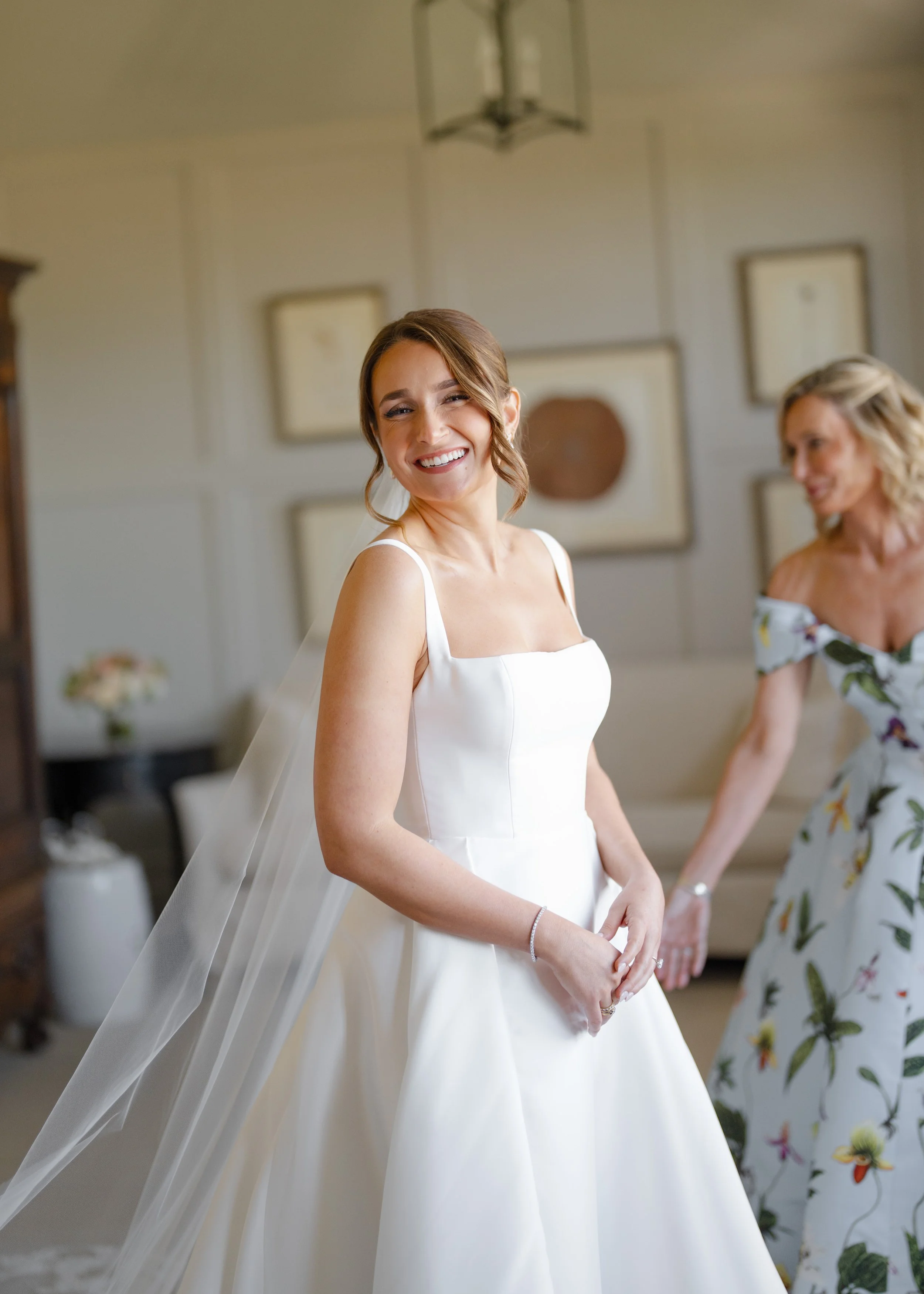 A smiling bride in a white wedding dress standing indoors, with a woman in a floral dress in the background.