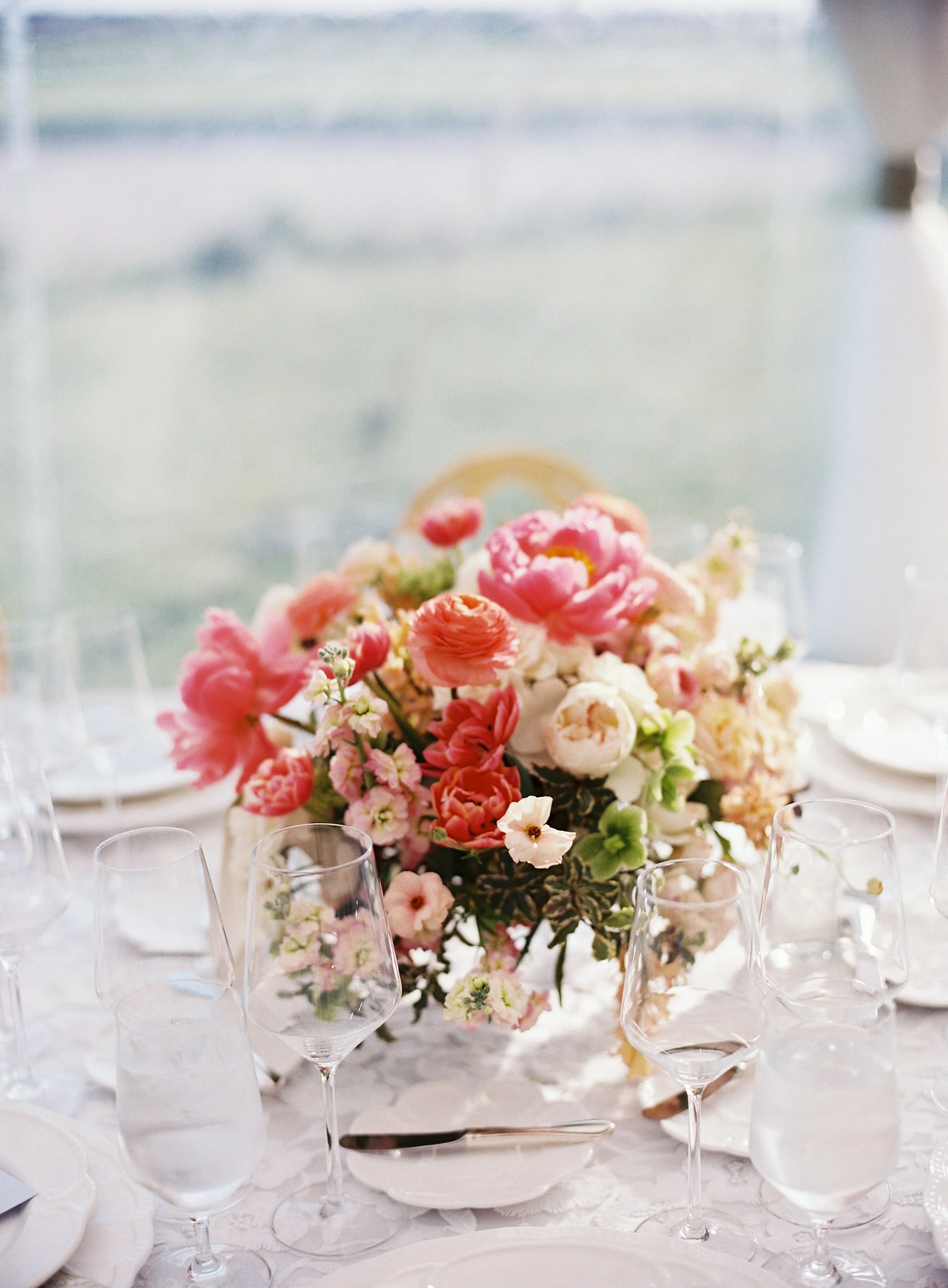 A wedding table centerpiece with pink and white flowers, surrounded by empty wine glasses and place settings on a white tablecloth.