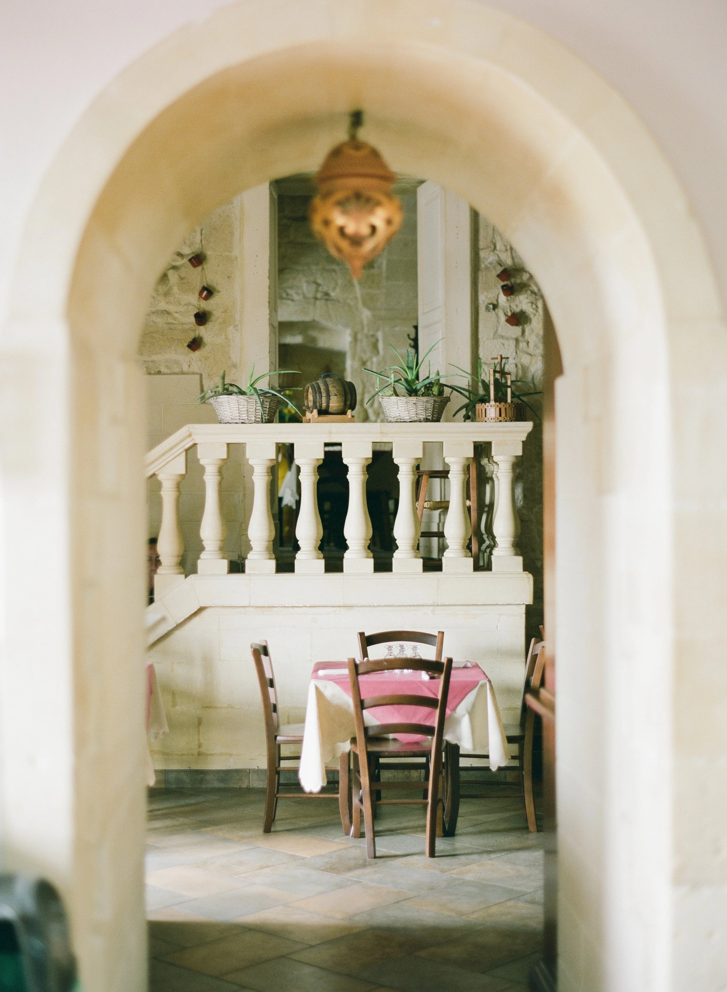 A cozy restaurant interior with a table covered with a pink and white tablecloth, surrounded by four wooden chairs, viewed through an arched doorway in Italy. 