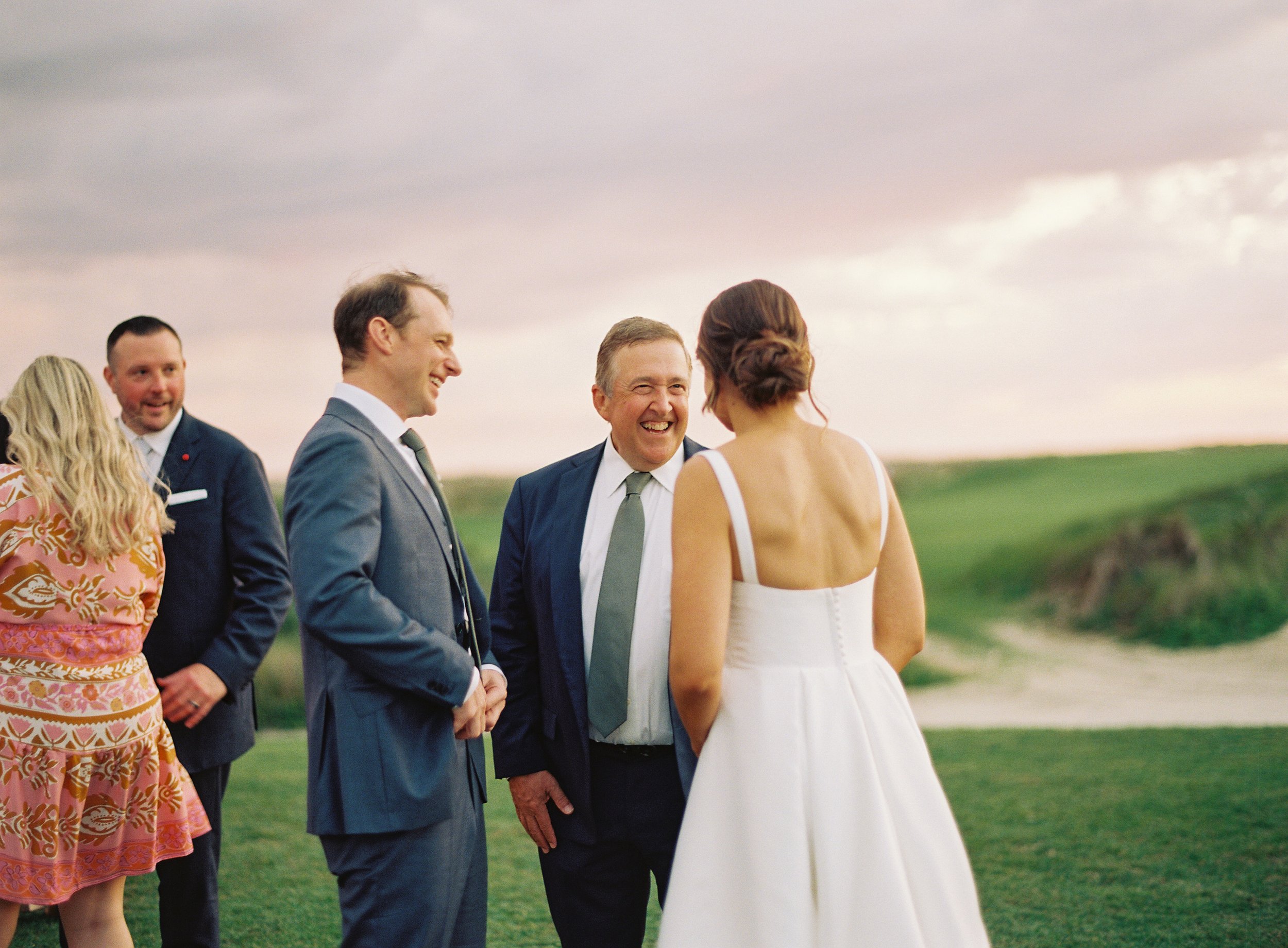 A bride in a white dress talking to groom and guests outdoors on a grassy field during sunset.