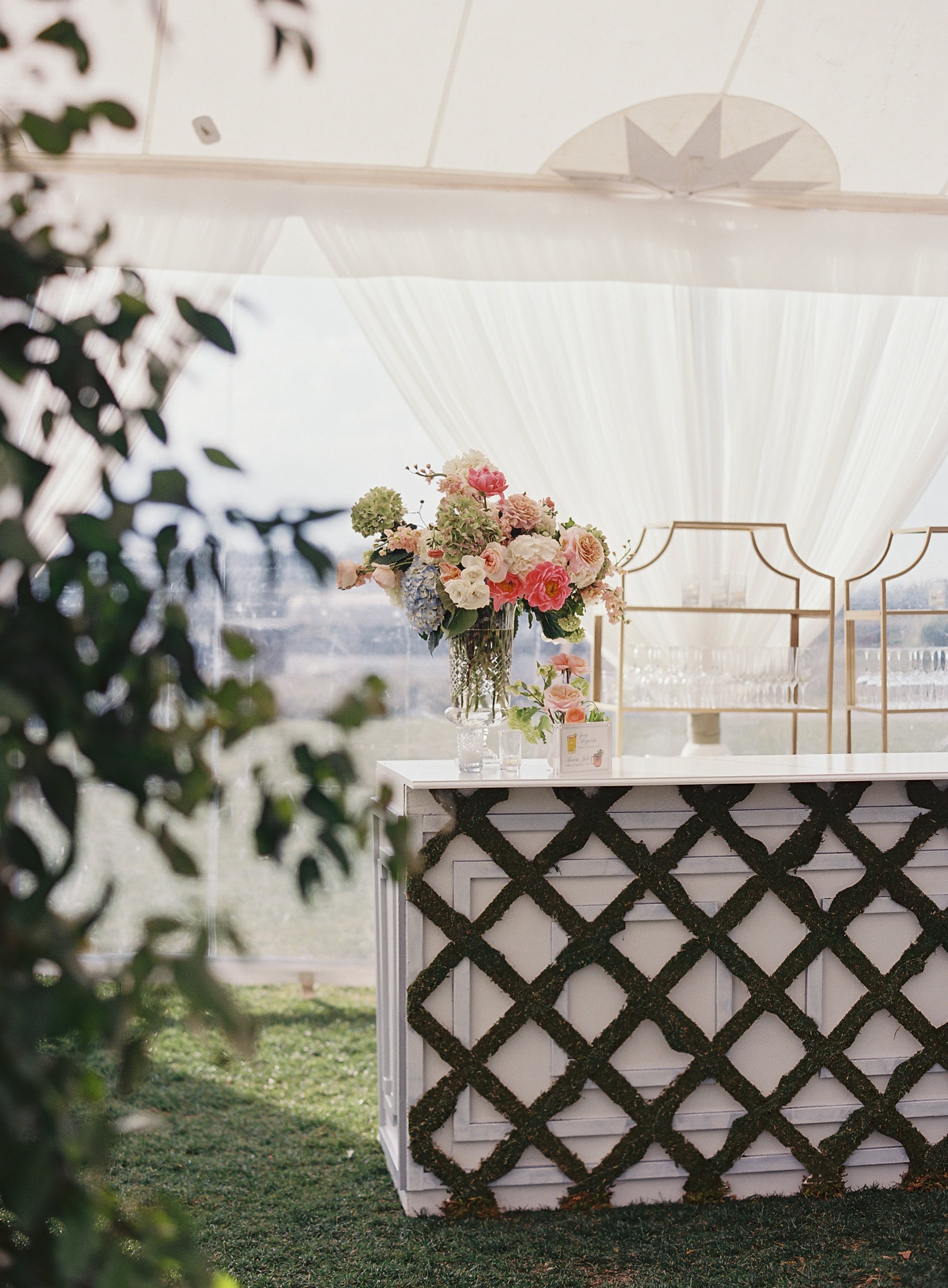 Beautiful flower arrangement on a white table at an outdoor event with a white tent and decorative gold accents.