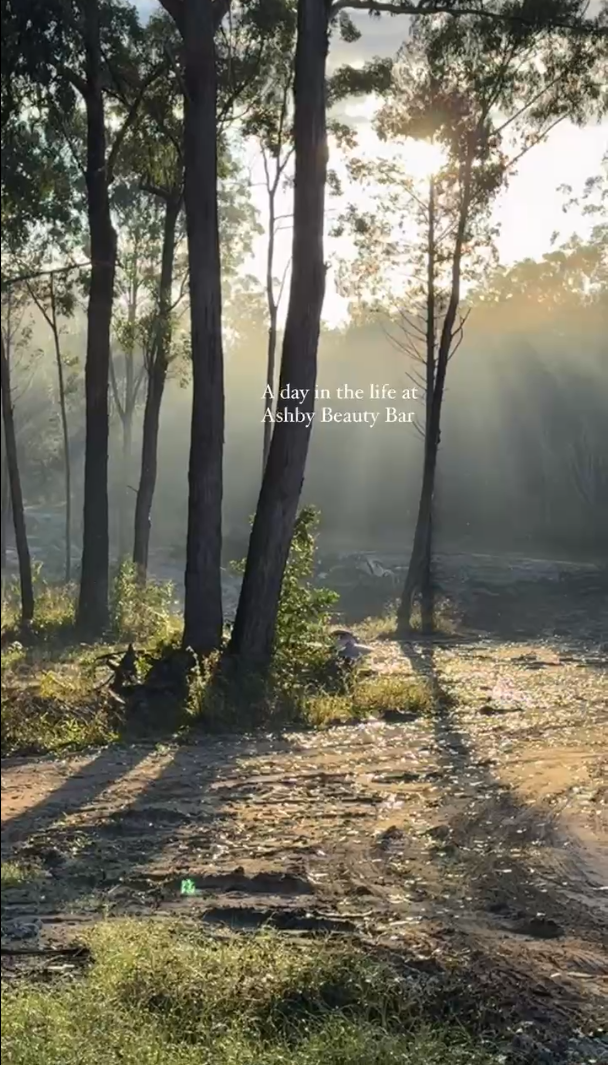 Sunlight shining through trees onto a dirt path in a forest with a caption reading "A day in the life at Ashby Beauty Bar".