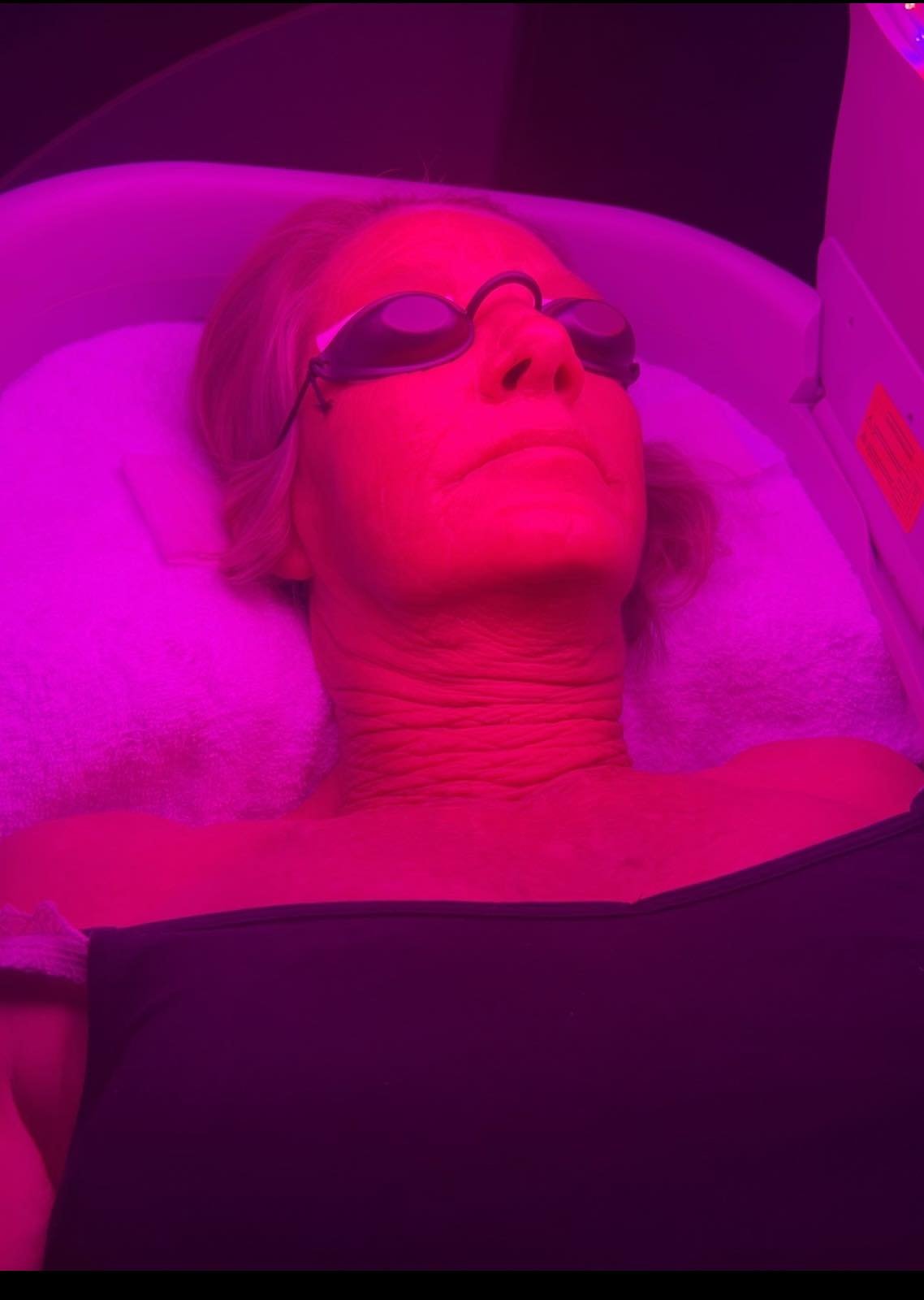 A woman lying under a Dermalux Flex with Red and NIR light, wearing black goggles, with her head resting on a pillow.