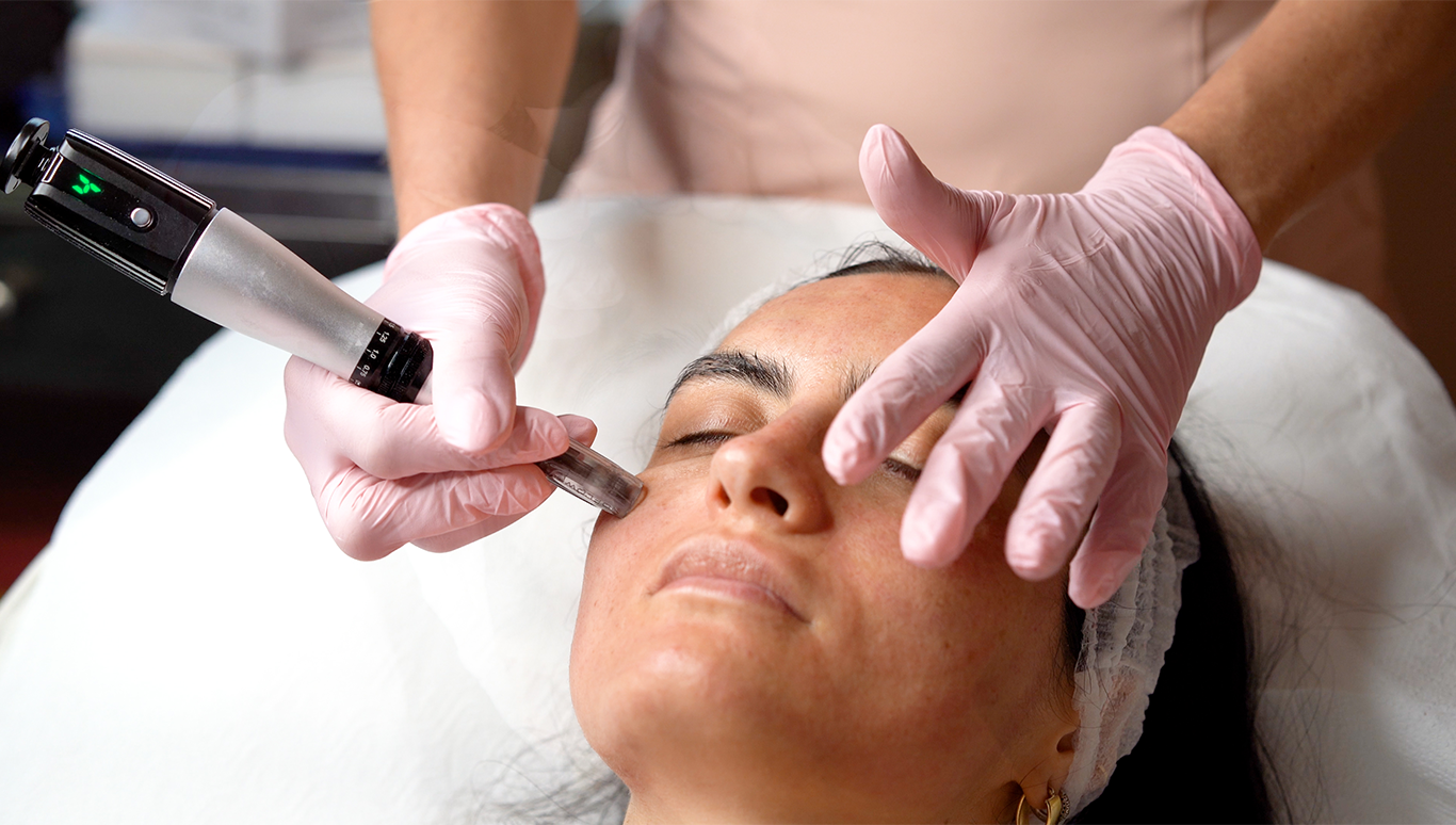 A person receiving a cosmetic facial treatment with a handheld device while lying down with a headband, wearing pink gloves.