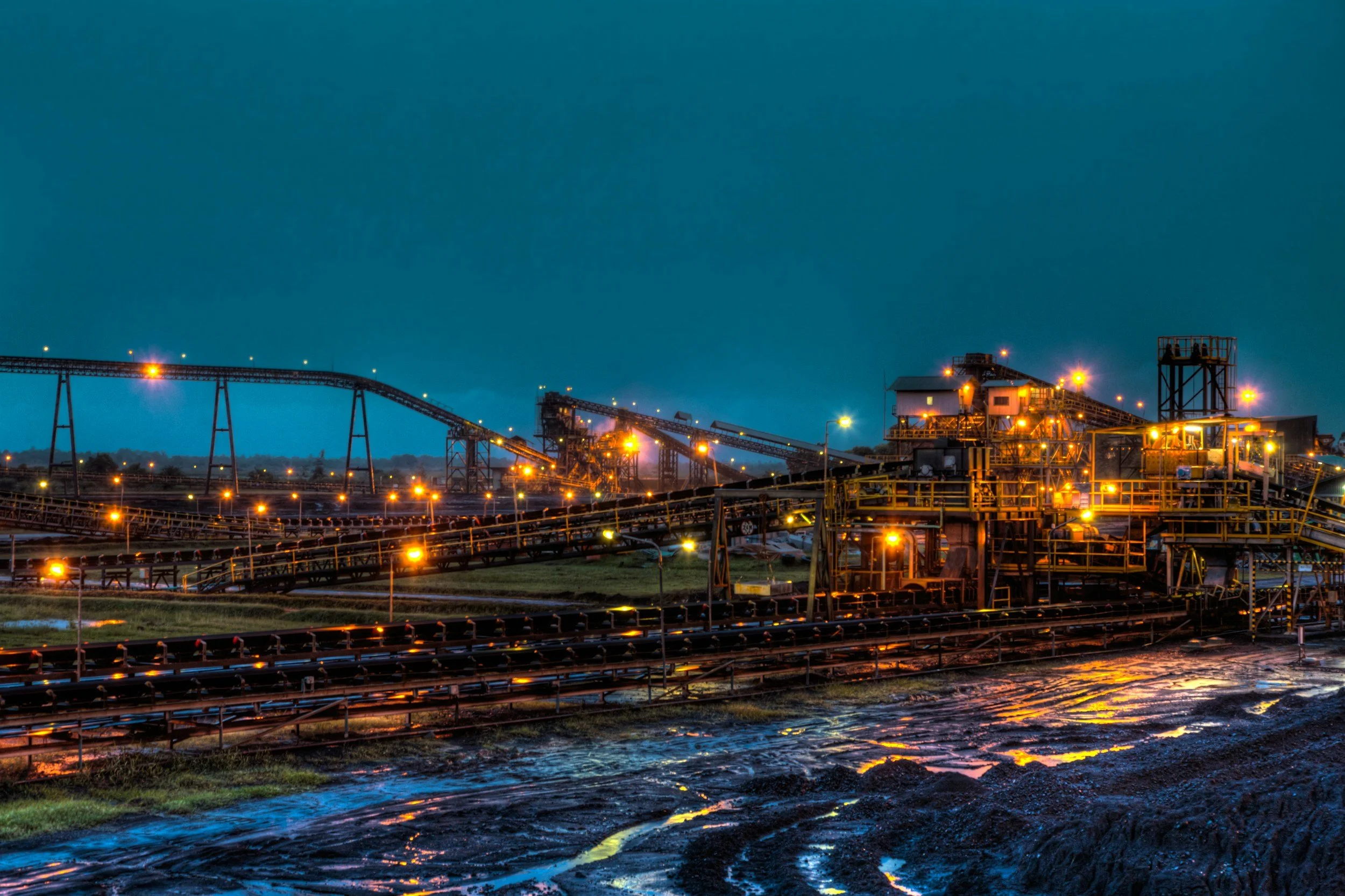 Industrial facility at night with conveyor belts and bright lights, reflections on wet ground.