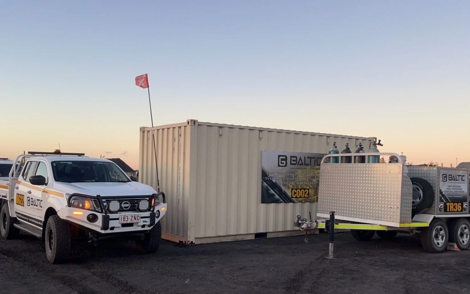 White utility vehicle and trailer next to a beige shipping container with signage from Baltic Projects, displaying gas cylinders on the trailer.