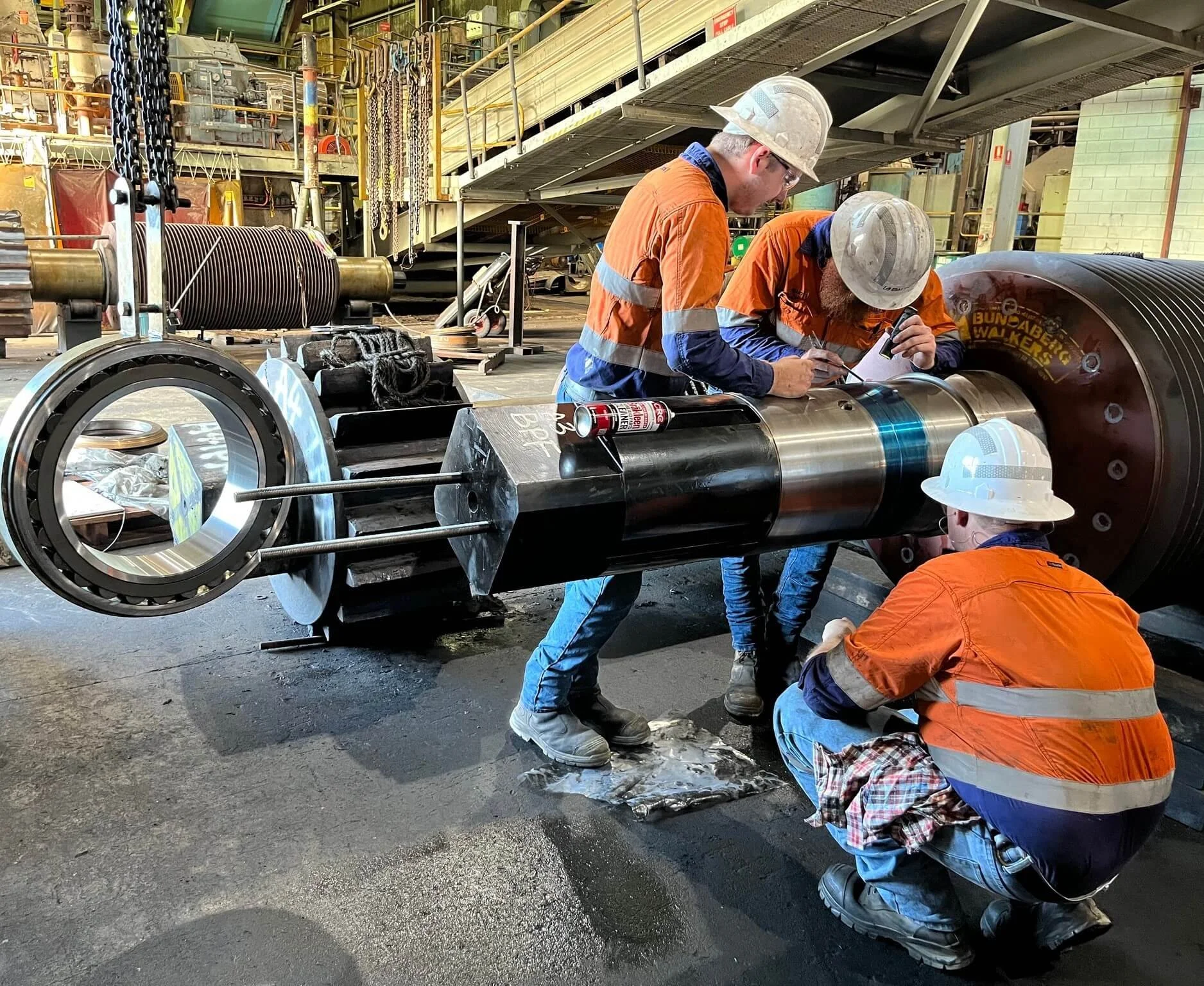Three workers in orange safety vests and helmets inspecting and working on a large industrial metal shaft in a warehouse setting.