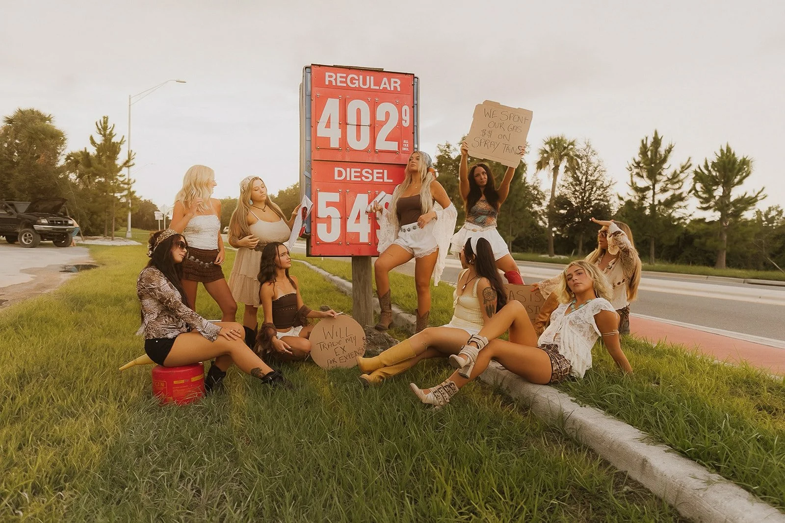 Group of young women protesting for climate change action on roadside with signs, sitting and standing near a gas price sign that reads 'Regular 402.9, Diesel 54.9'.