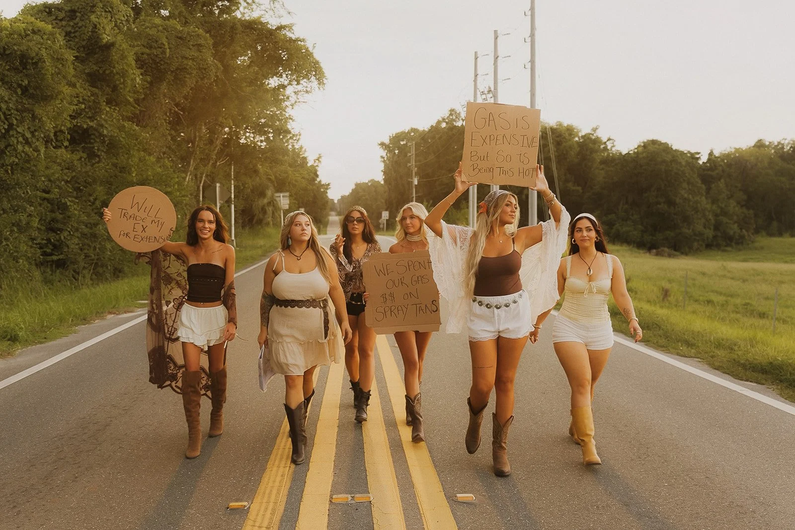 Group of women protesting on a country road holding signs about gas prices and credit issues. They are walking across the street with trees and power lines in the background.