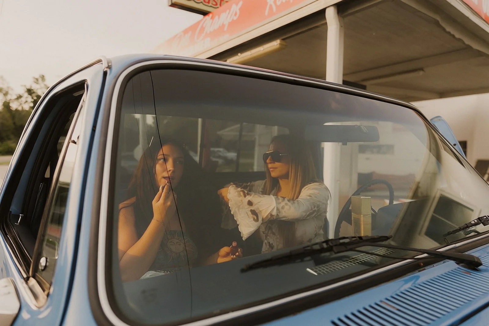 Two women sitting inside a vintage car, one is applying lipstick and the other is pointing at her, with a background of a gas station or roadside stop.