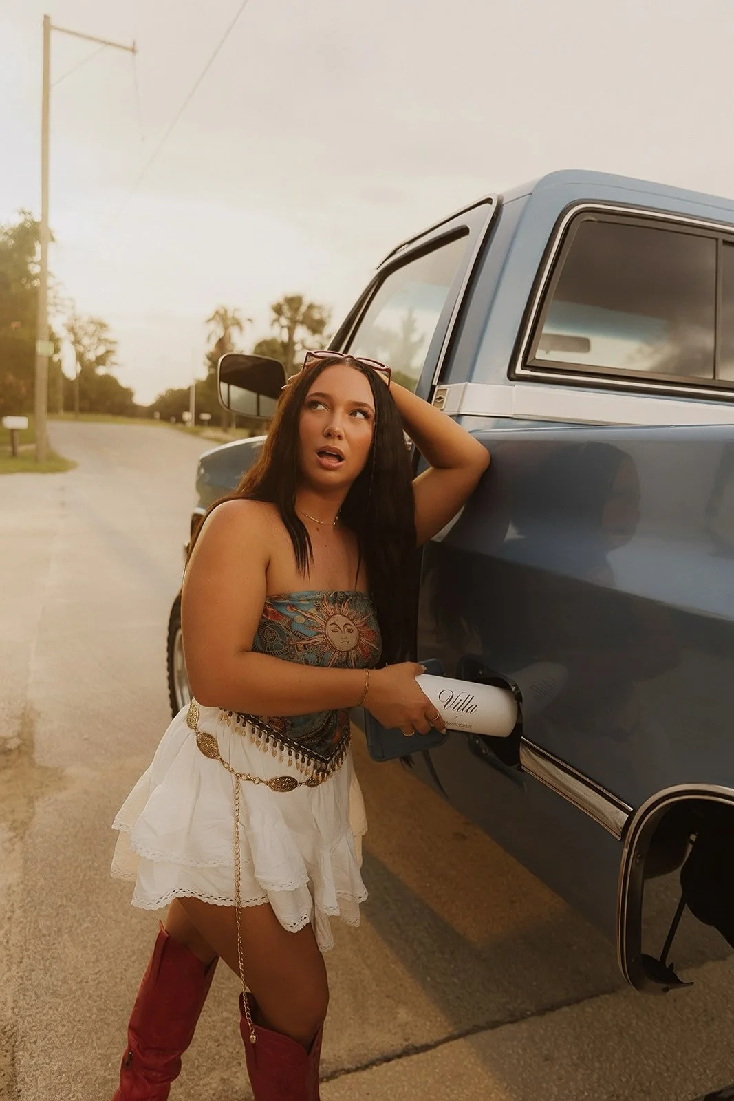 A woman in a strapless top and white skirt stands next to a blue pickup truck on a rural road during sunset, holding a white water bottle with the word 'Villa' on it.