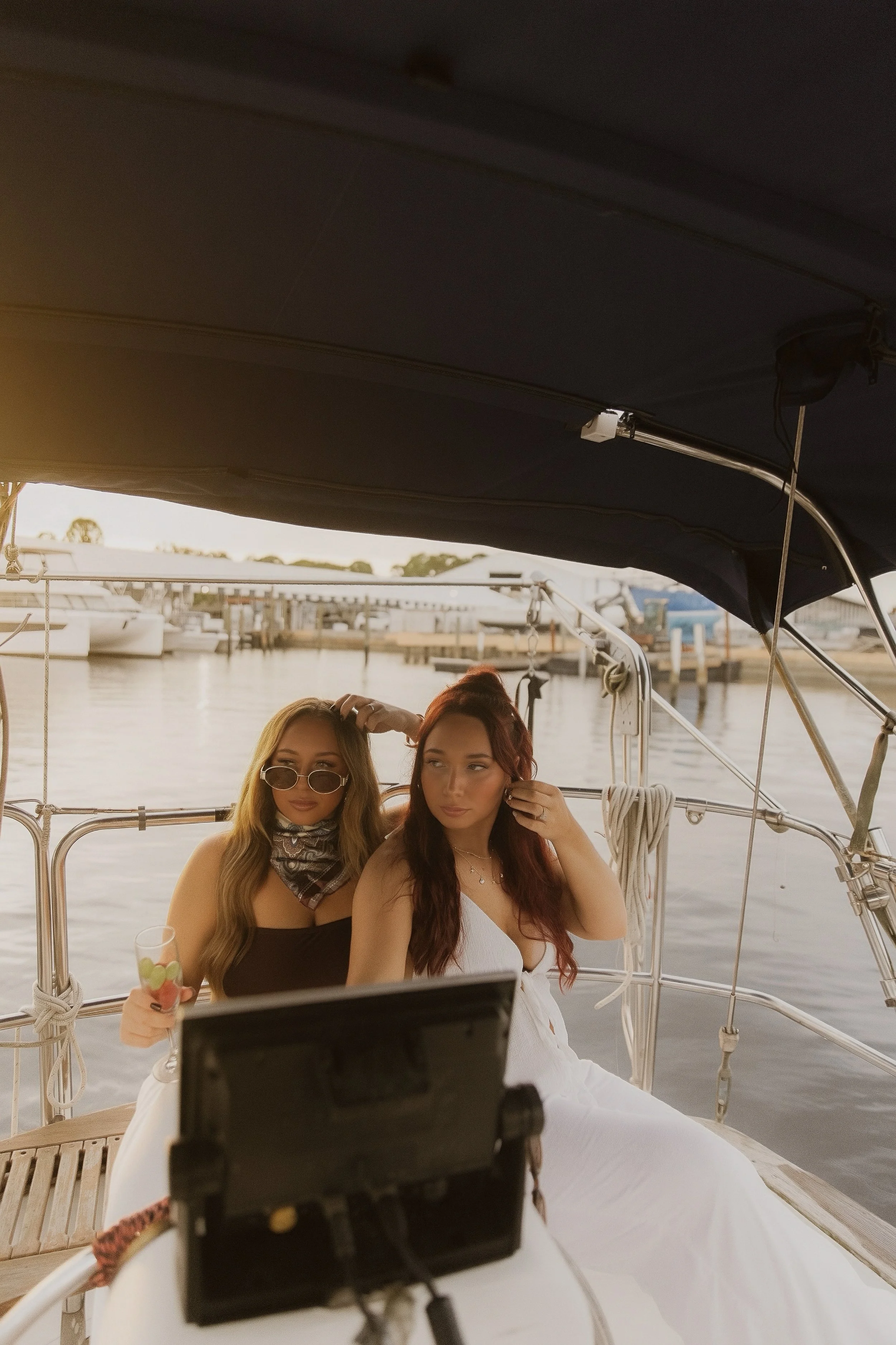 Two young women sitting on a boat docked at a marina, taking a selfie with a camera mounted on a tripod in front of them. The woman on the left wears sunglasses and a bandana, holding a drink, while the woman on the right has long red hair and is wearing a white dress. There are boats and the water in the background.