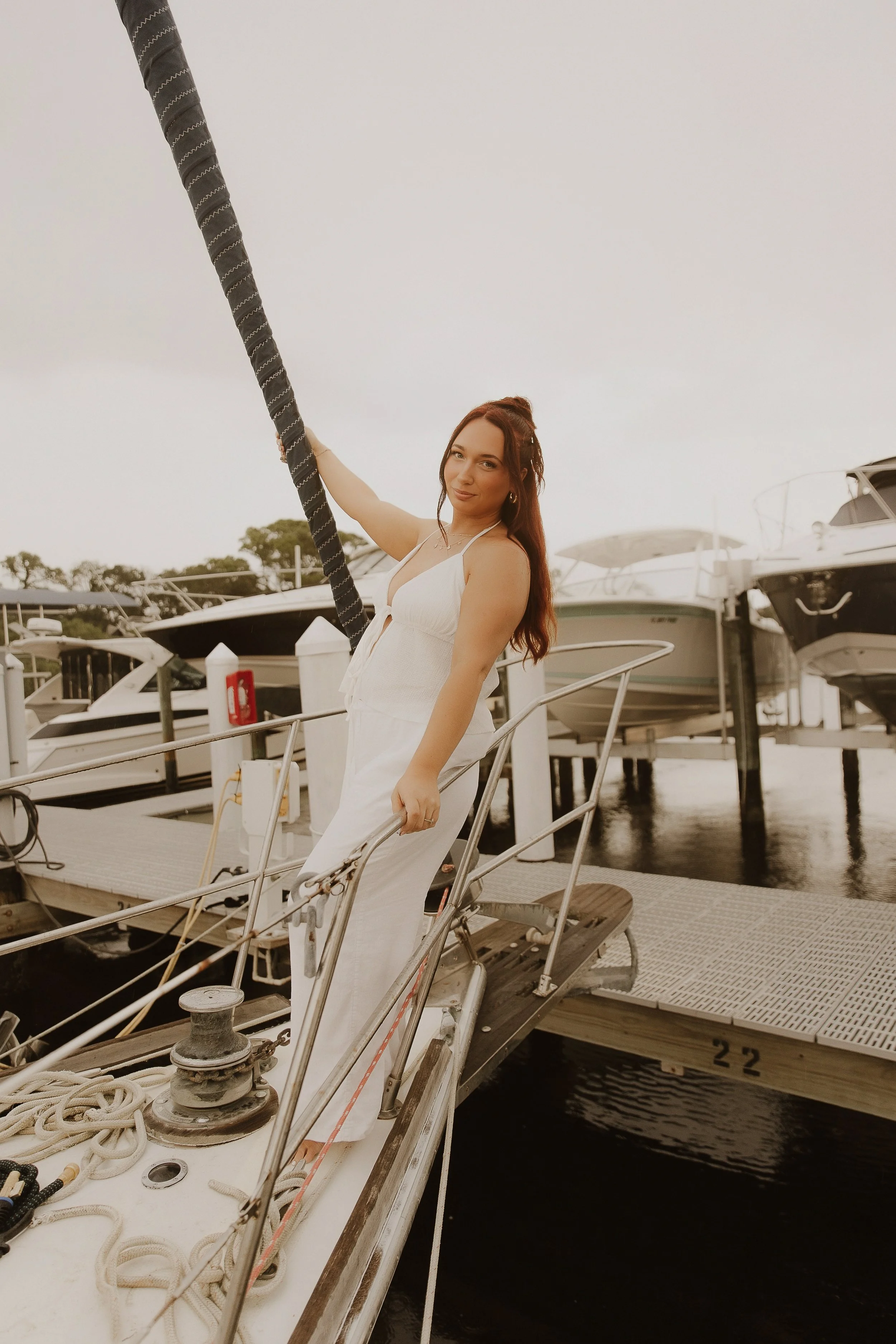 Woman with red hair in a white sleeveless dress standing on a boat dock, holding a rope, with several boats in the background.