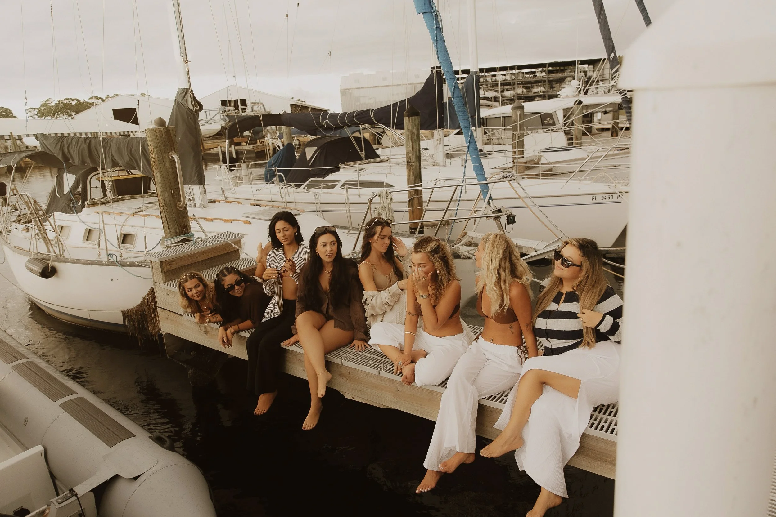 A group of women sitting on a dock with their feet dangling over the edge, near sailboats docked at a marina.