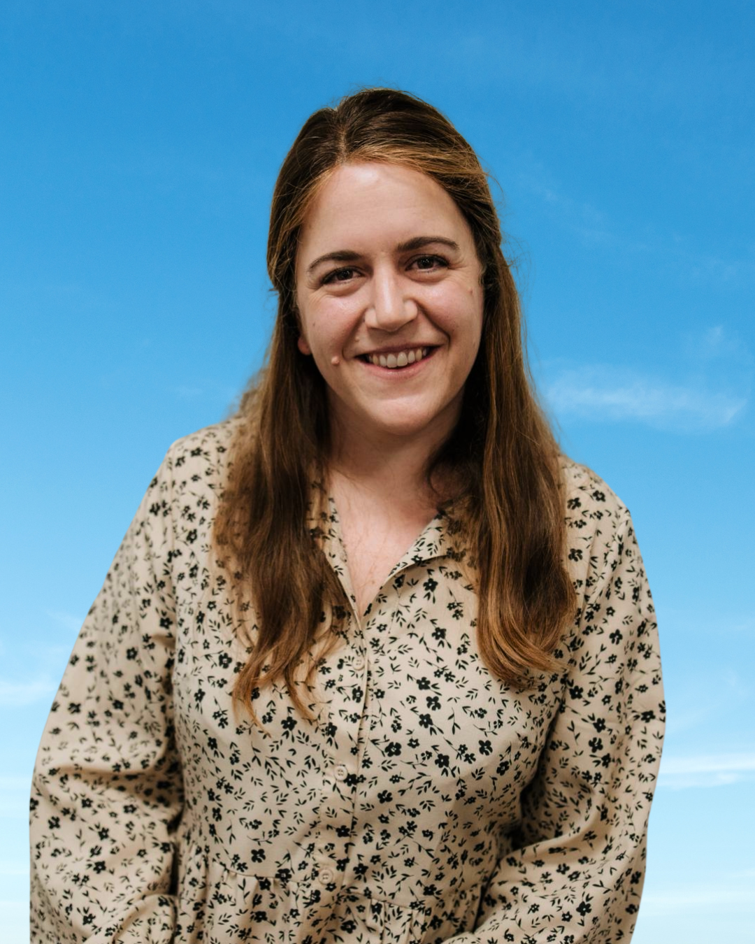 A woman with brown hair smiling, wearing a beige shirt with black floral patterns, standing against a blue sky background.