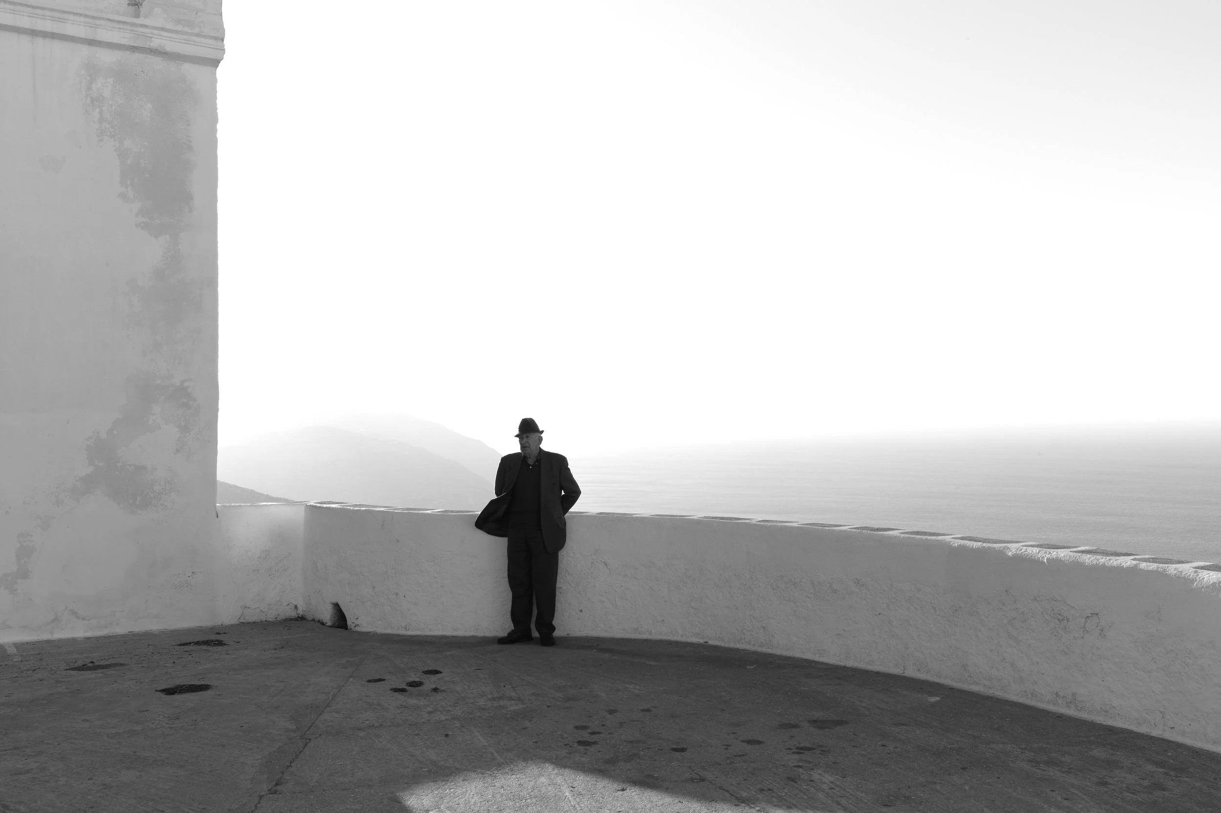 Schwarz-weiß-Foto eines Mannes, der auf einer Terrasse mit Blick auf das Meer steht, mit einem Berg im Hintergrund. Der Mann trägt einen Hut und einen Anzug.
