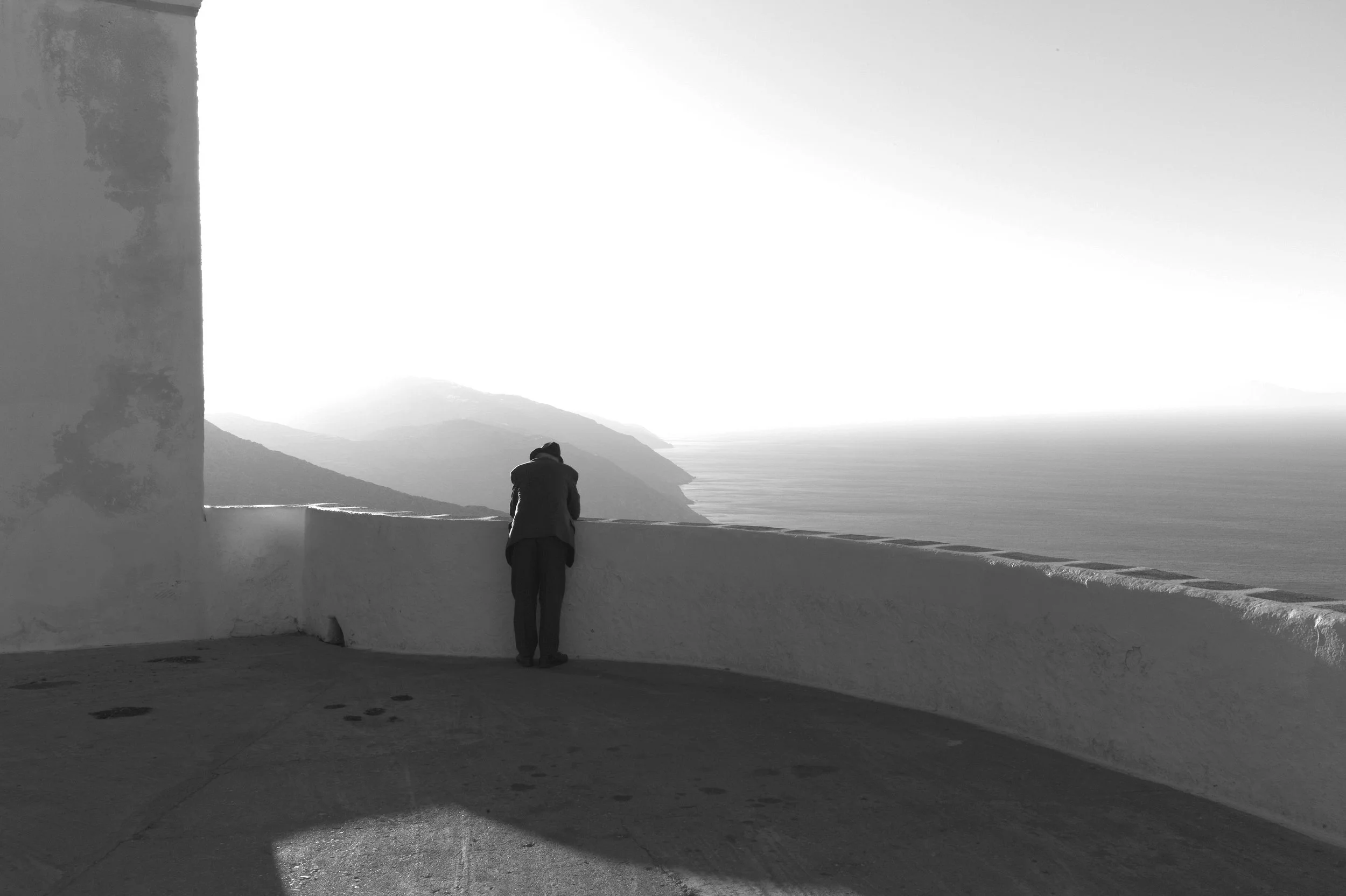 Ein Mensch lehnt auf einer weißen Mauer mit Blick auf das Meer und die Berge im Hintergrund. Es ist eine Schwarz-Weiß-Fotografie.