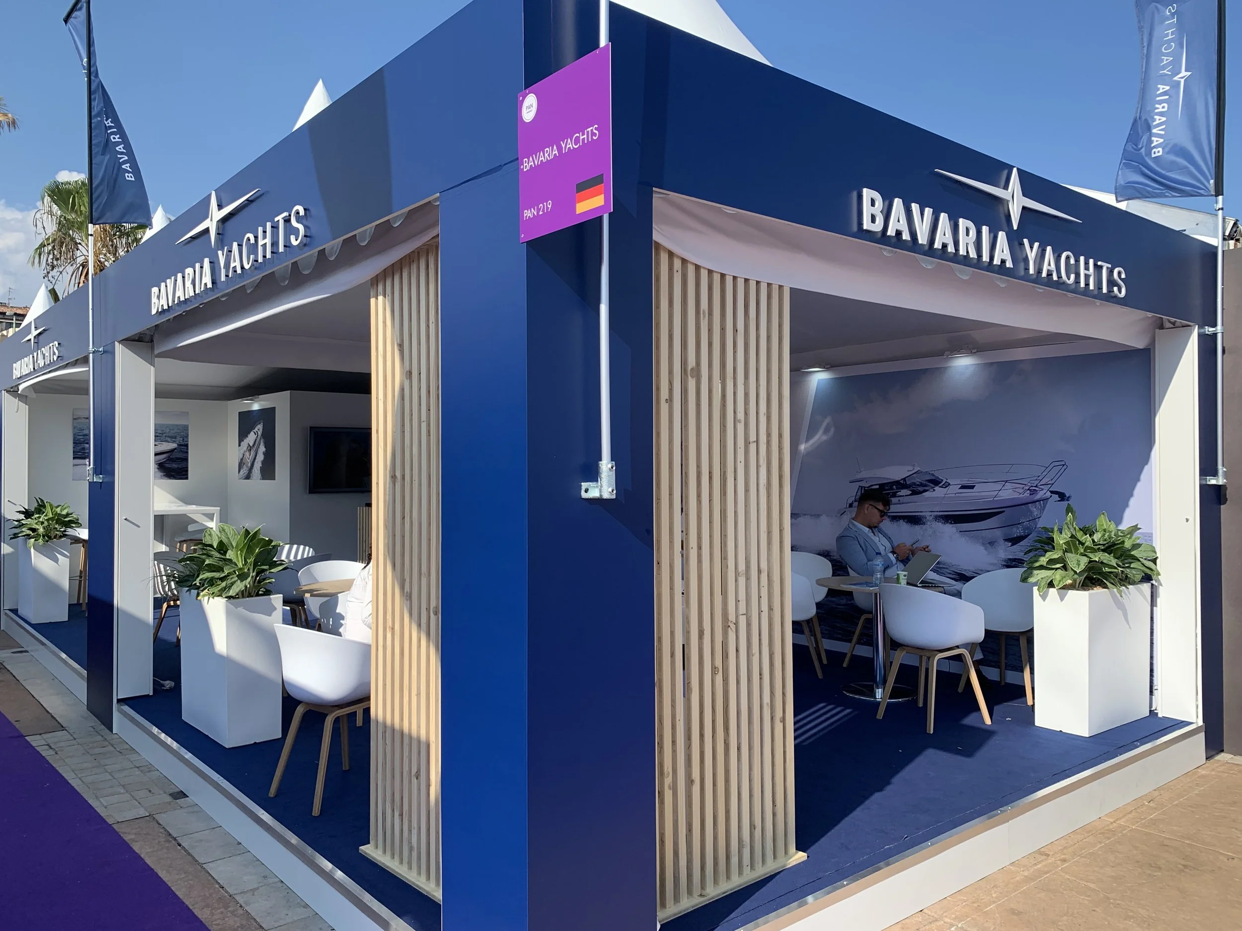 Stand Bavaria Yachts avec décoration nautique, plantes vertes, et un homme utilisant un ordinateur portable à l'intérieur.