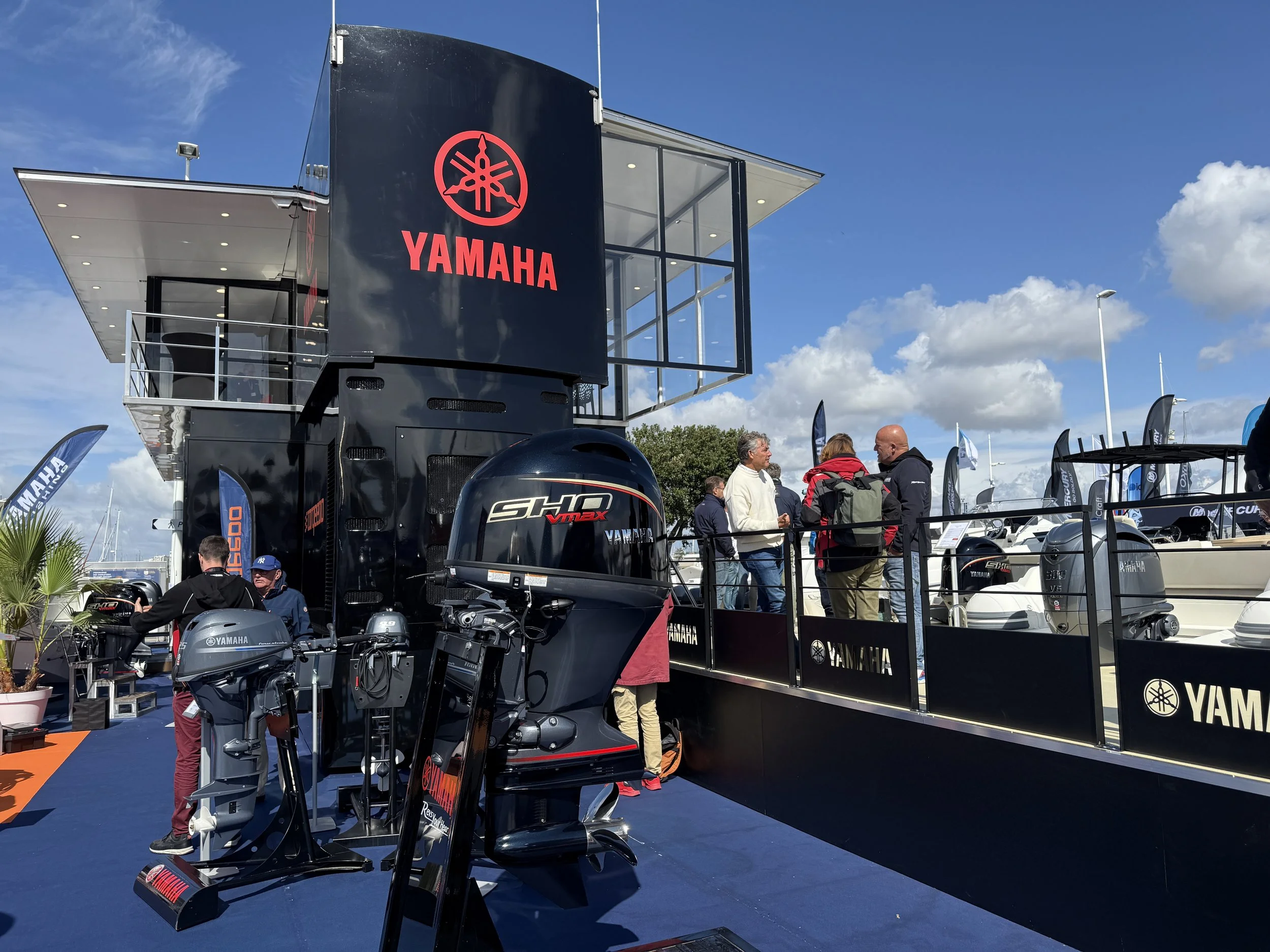Exposition Yamaha avec moteurs hors-bord et moteurs de bateau, plusieurs personnes discutant, bateau visible à côté.