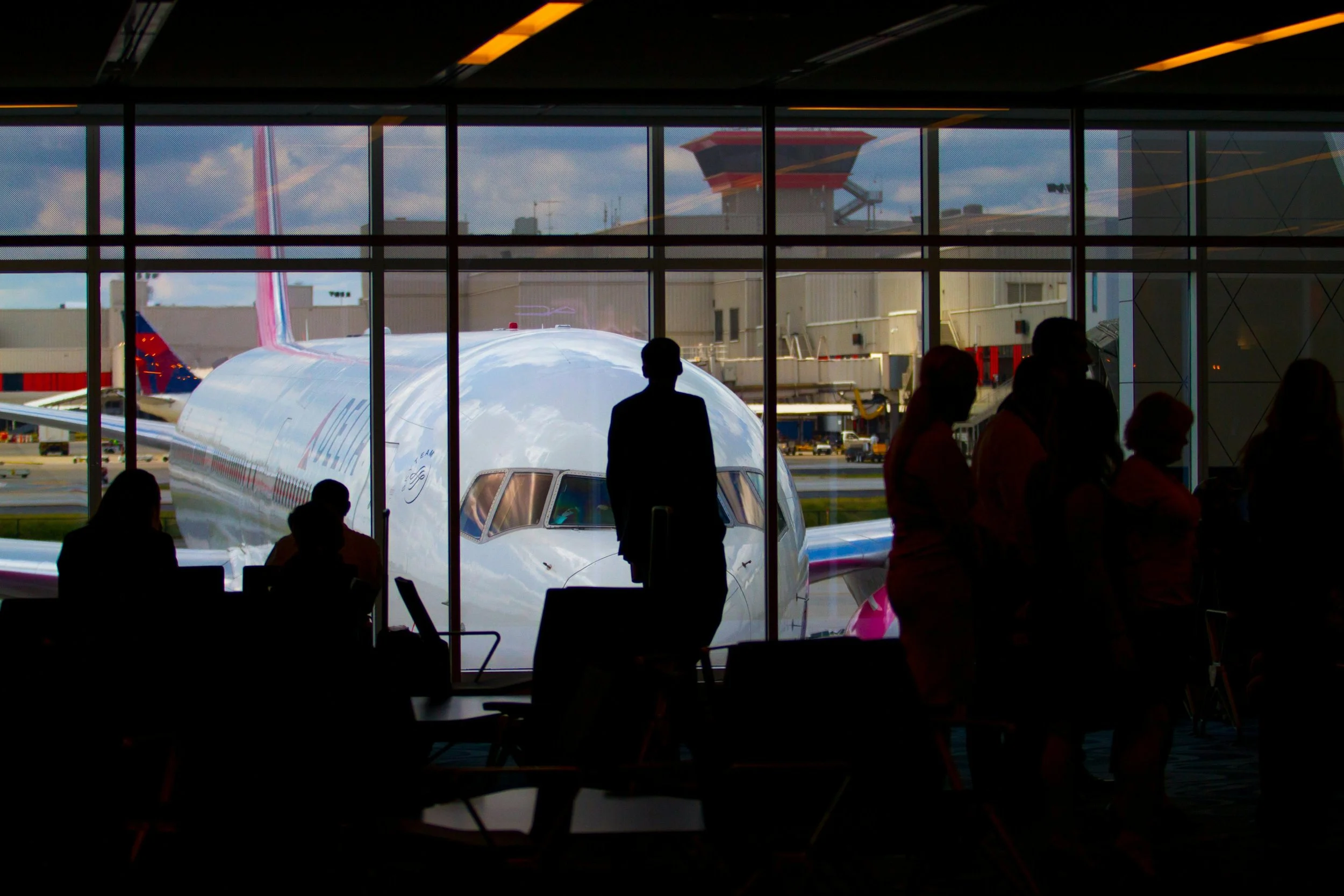 Regard à travers une grande fenêtre d'aéroclub, avec des silhouettes de passagers assis et debout à l'intérieur, un avions de ligne blanc en marche à l’extérieur, avec un terminal et des nuages en arrière-plan.